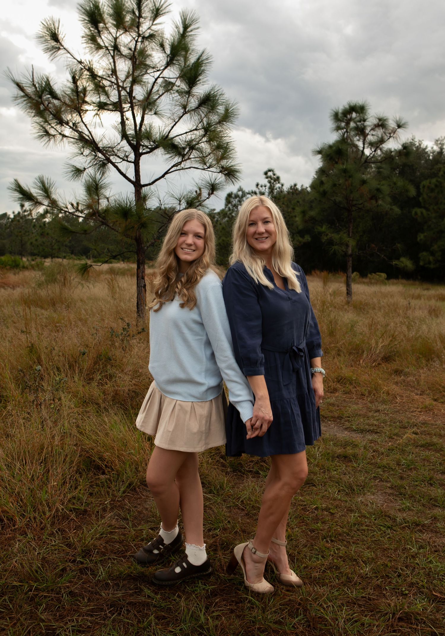 Two smiling women hold hands while standing in a grassy field beside a young pine tree.