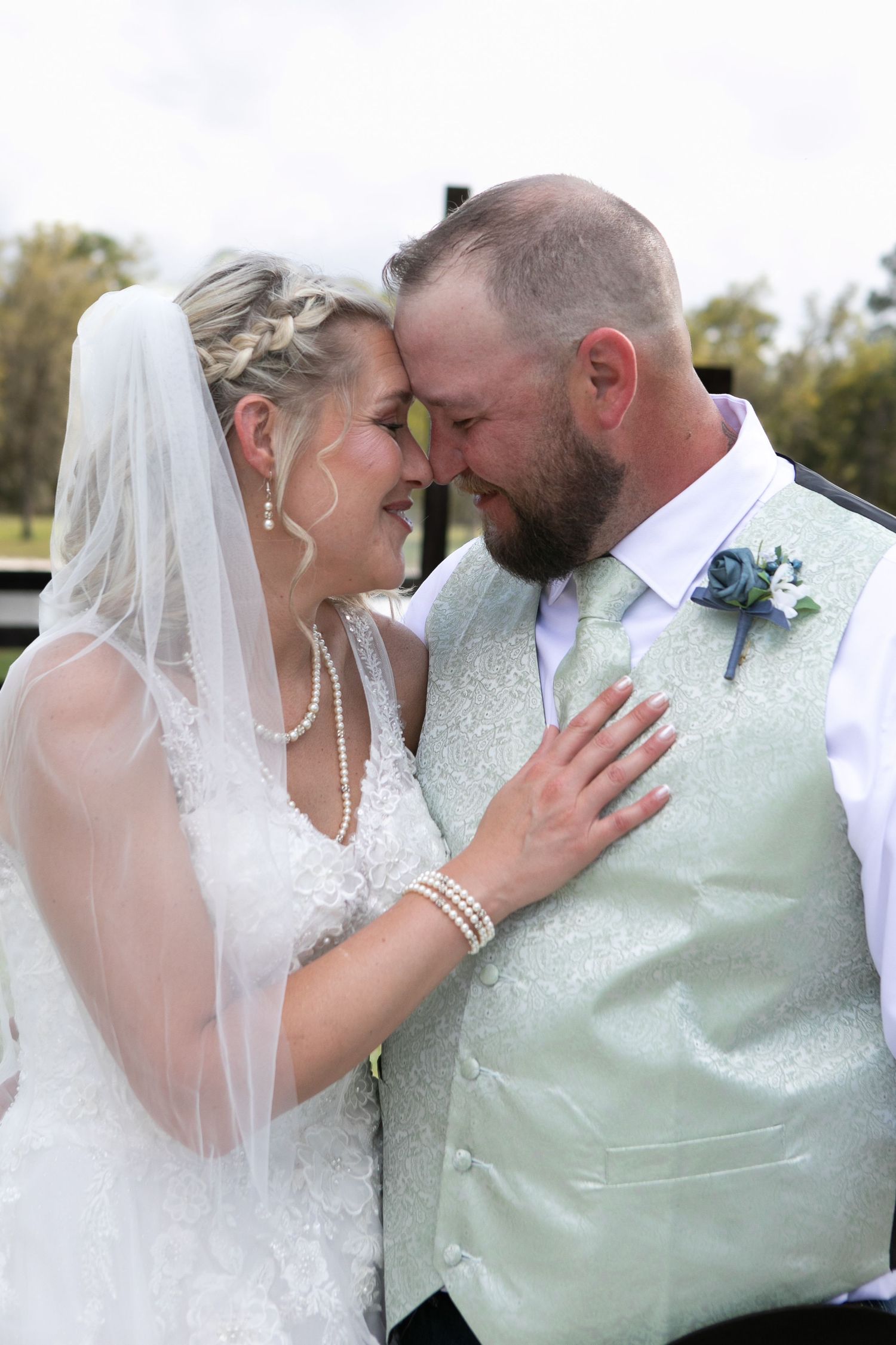 Bride and bearded groom touch foreheads tenderly outdoors, she wears a veil and pearl jewelry on their wedding day.