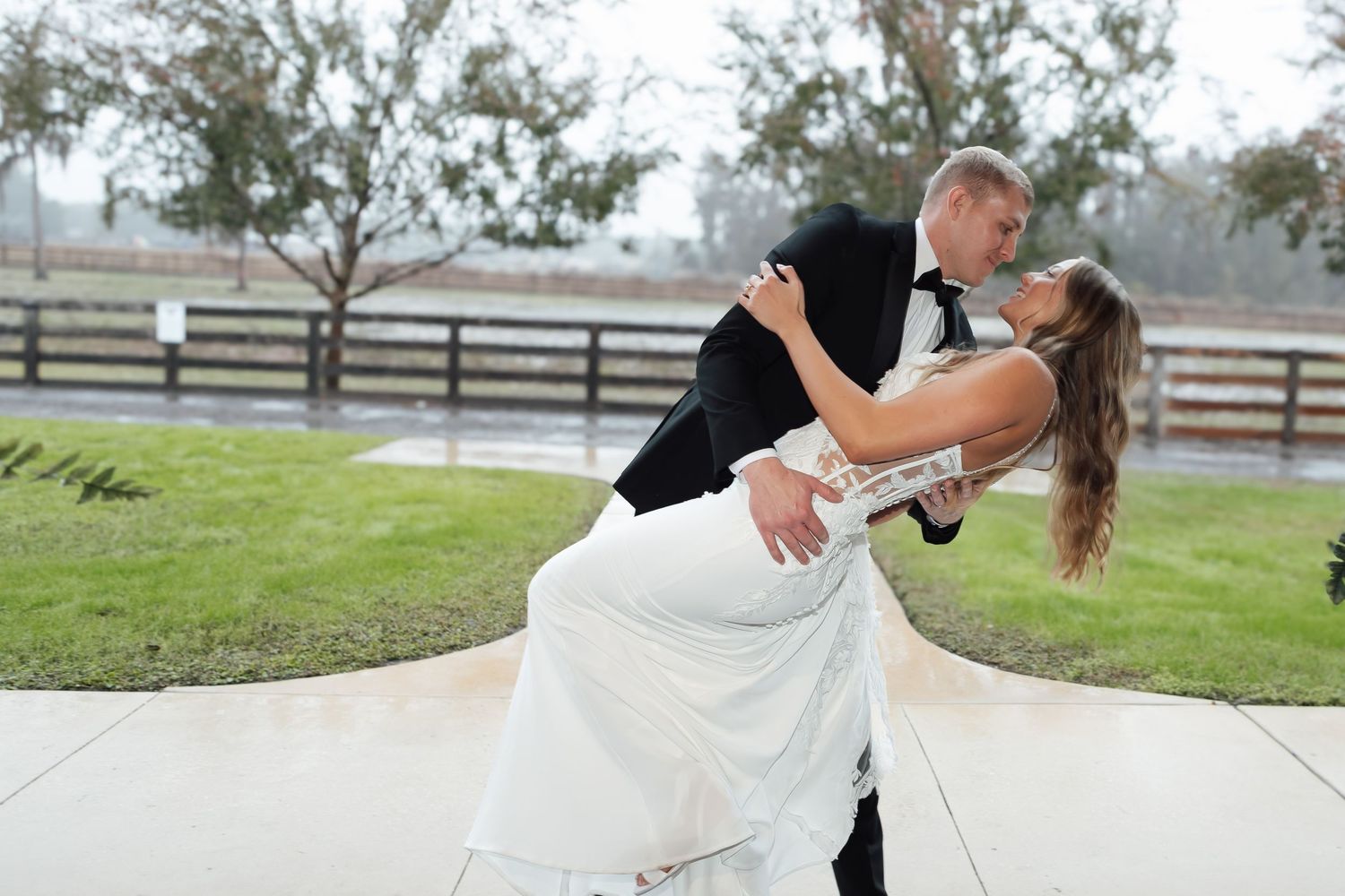 Groom dips bride in white wedding dress during outdoor dance at rustic venue.