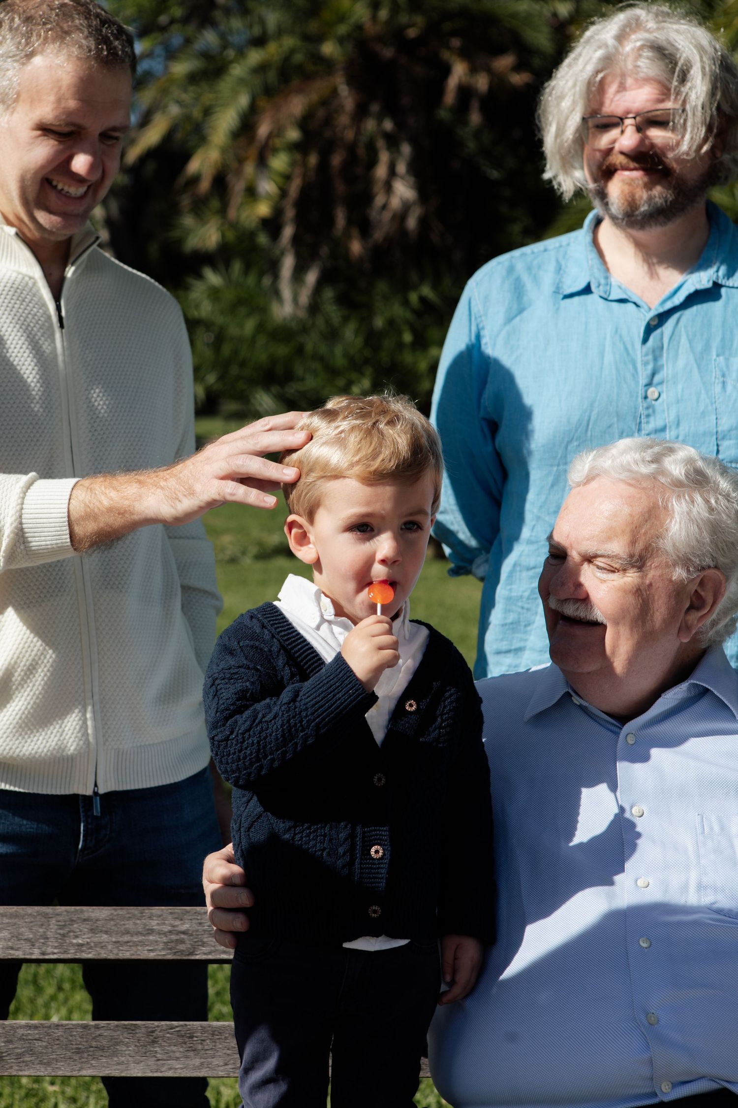 Multi-generational family portrait outdoors with grandfather, father, and toddler eating a popsicle in a sunny park.