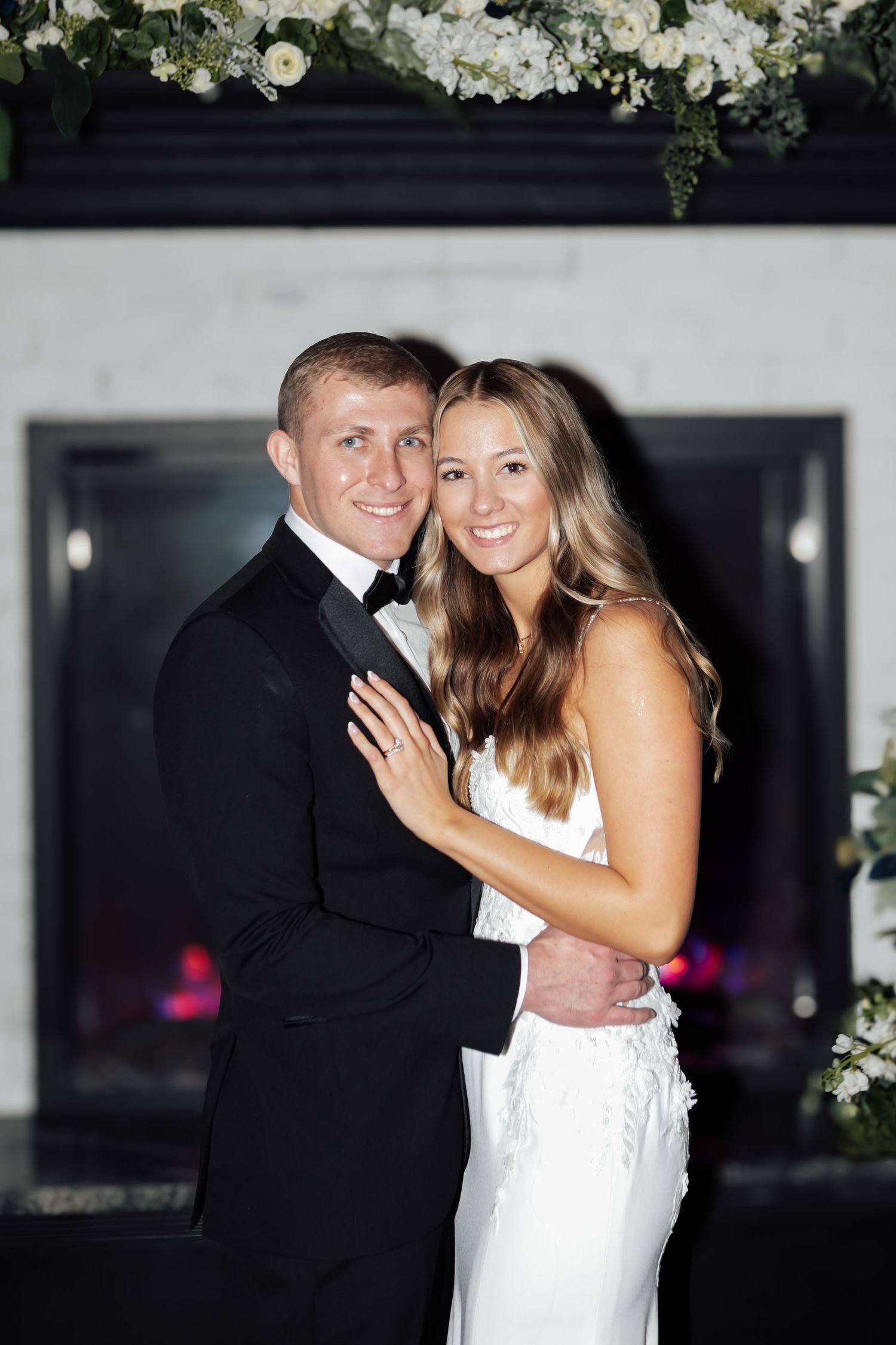 Newlywed couple poses elegantly in front of a floral-adorned fireplace at their wedding reception.