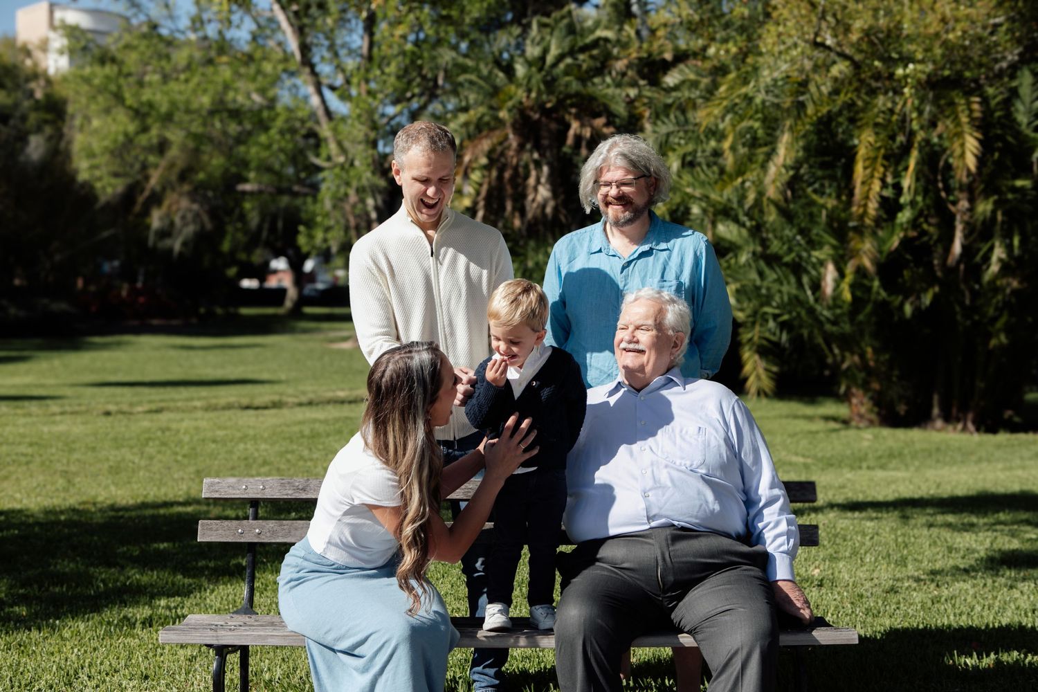 Multi-generational family of five gathers on a park bench in a lush green outdoor setting on a sunny day.