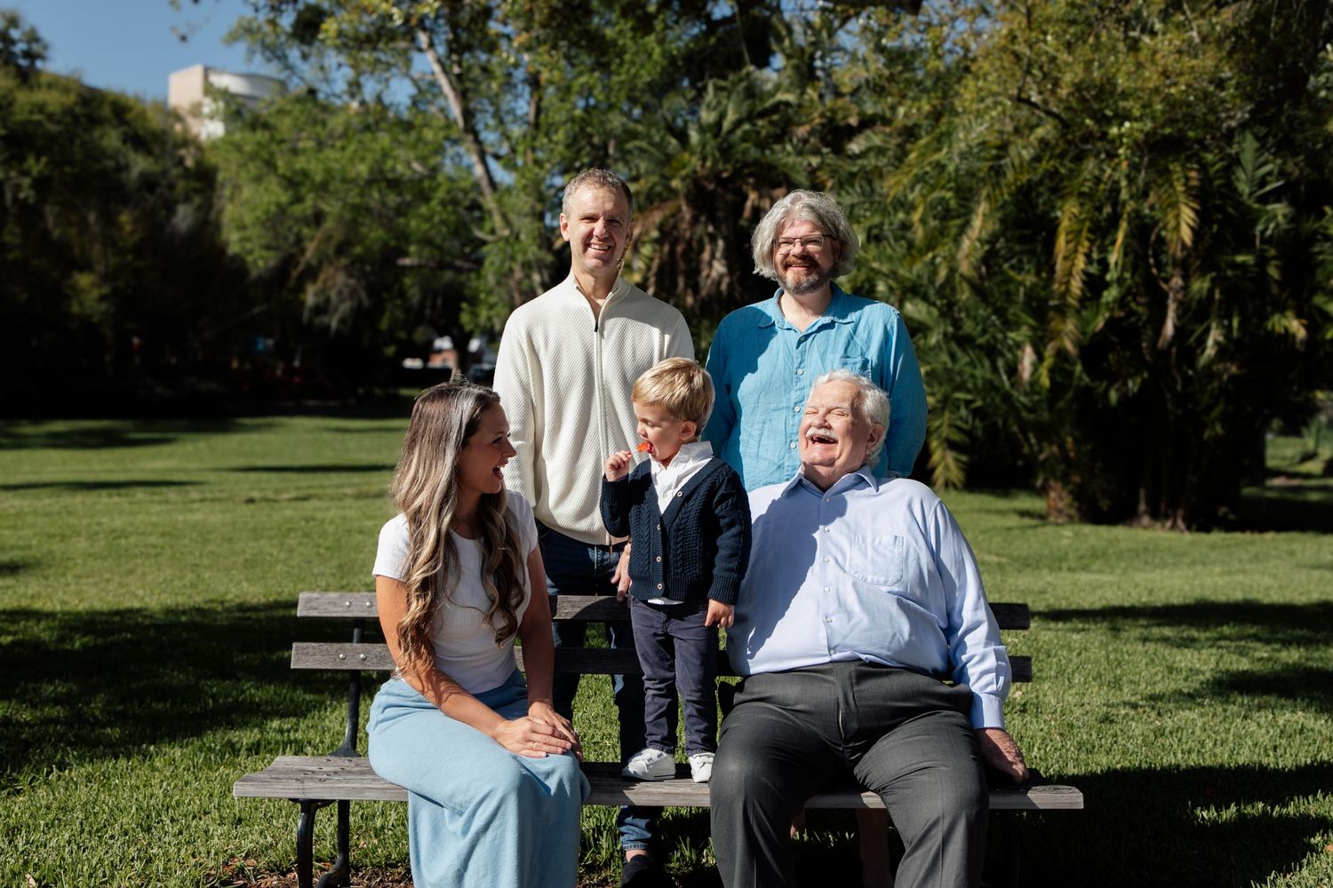 Multi-generational family smiling together on park benches surrounded by lush green trees on a sunny day.