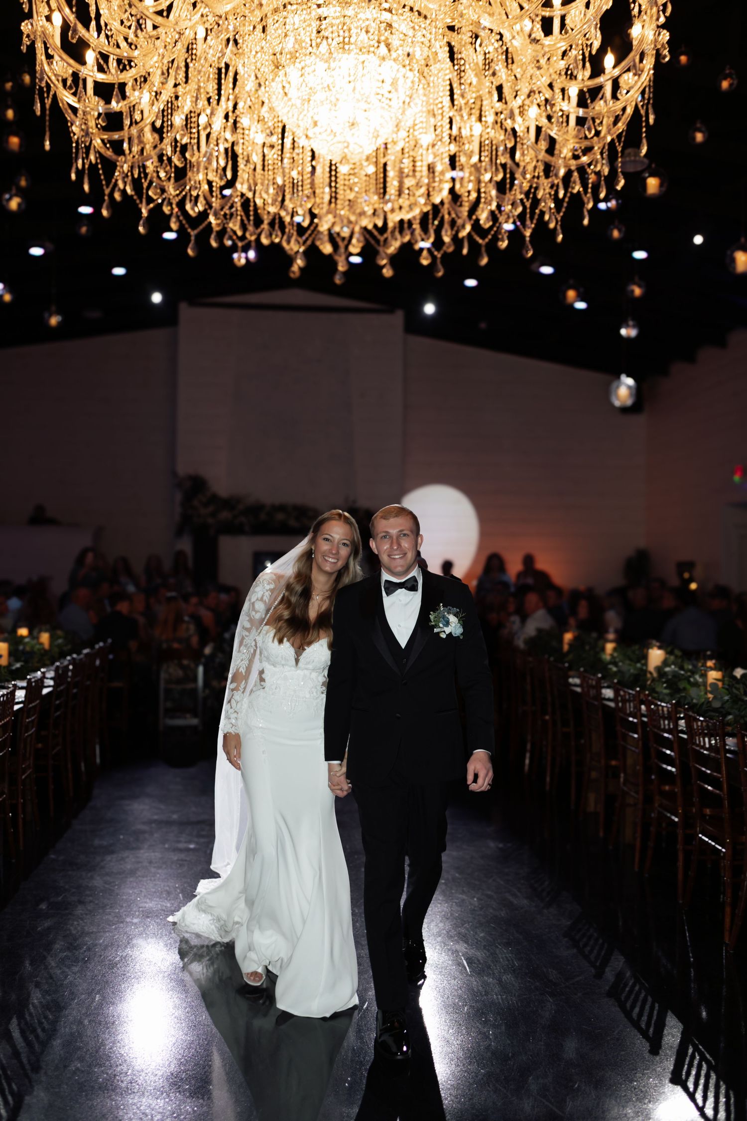 Bride and groom walking down the aisle under a grand crystal chandelier at their reception.