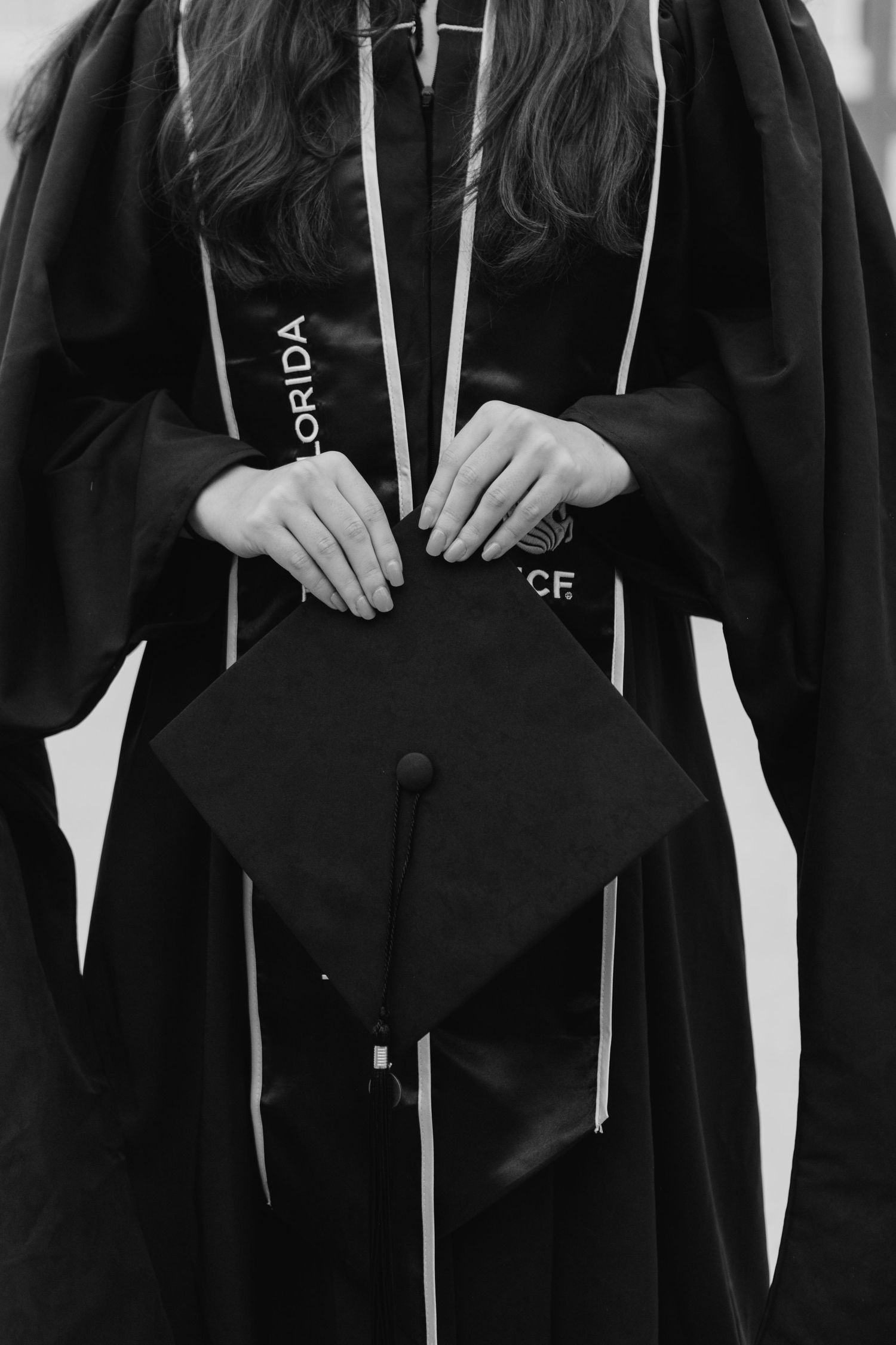 Close-up black and white photo of a graduate holding mortarboard cap wearing Florida UCF gown.