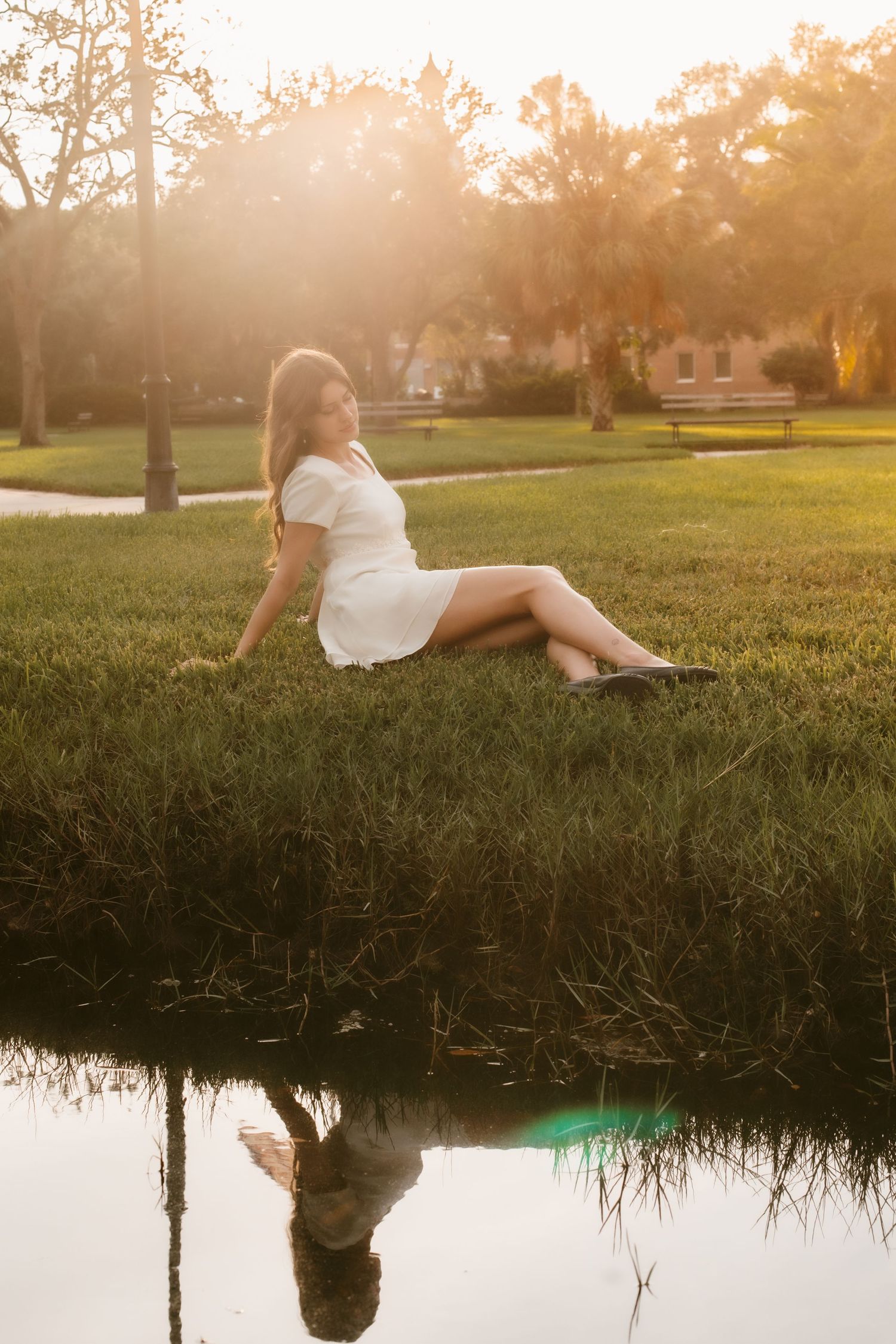 Young woman in white dress sitting on lush green grass by a reflective pond at golden hour sunset.