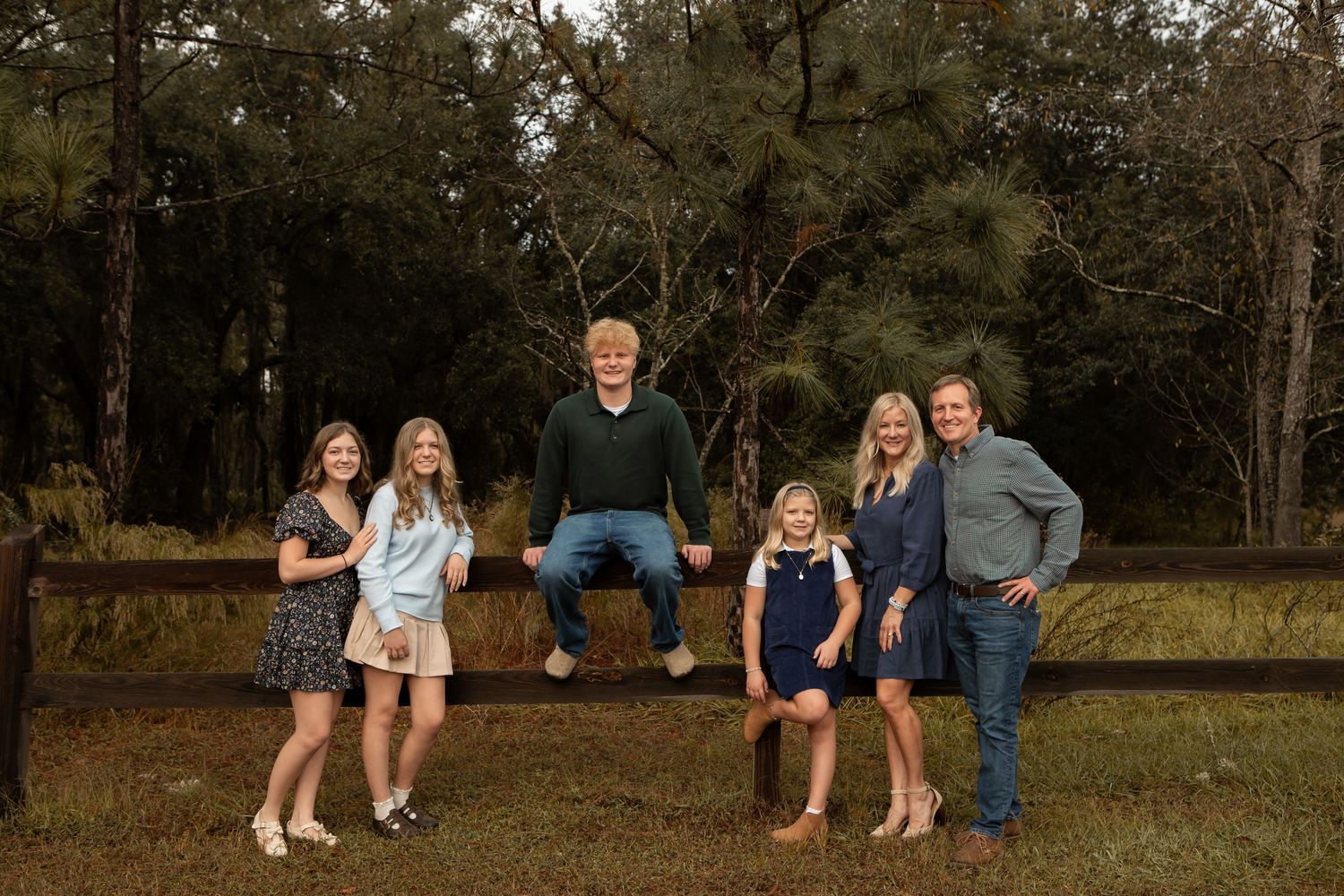 Family of six smiling together outdoors by a wooden fence surrounded by lush green trees.