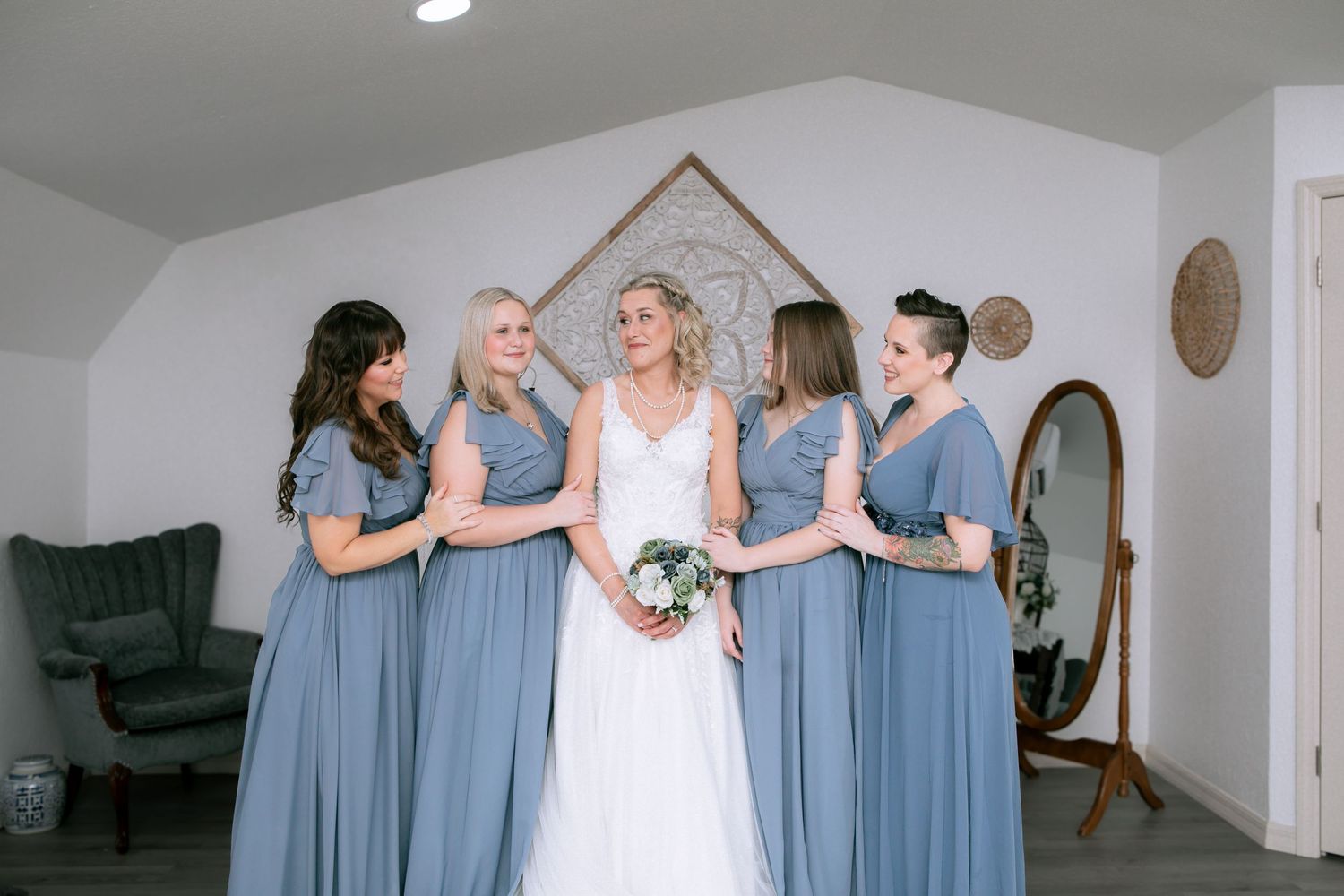 Bride in white gown stands with four bridesmaids in dusty blue dresses, smiling together in a bright indoor setting.