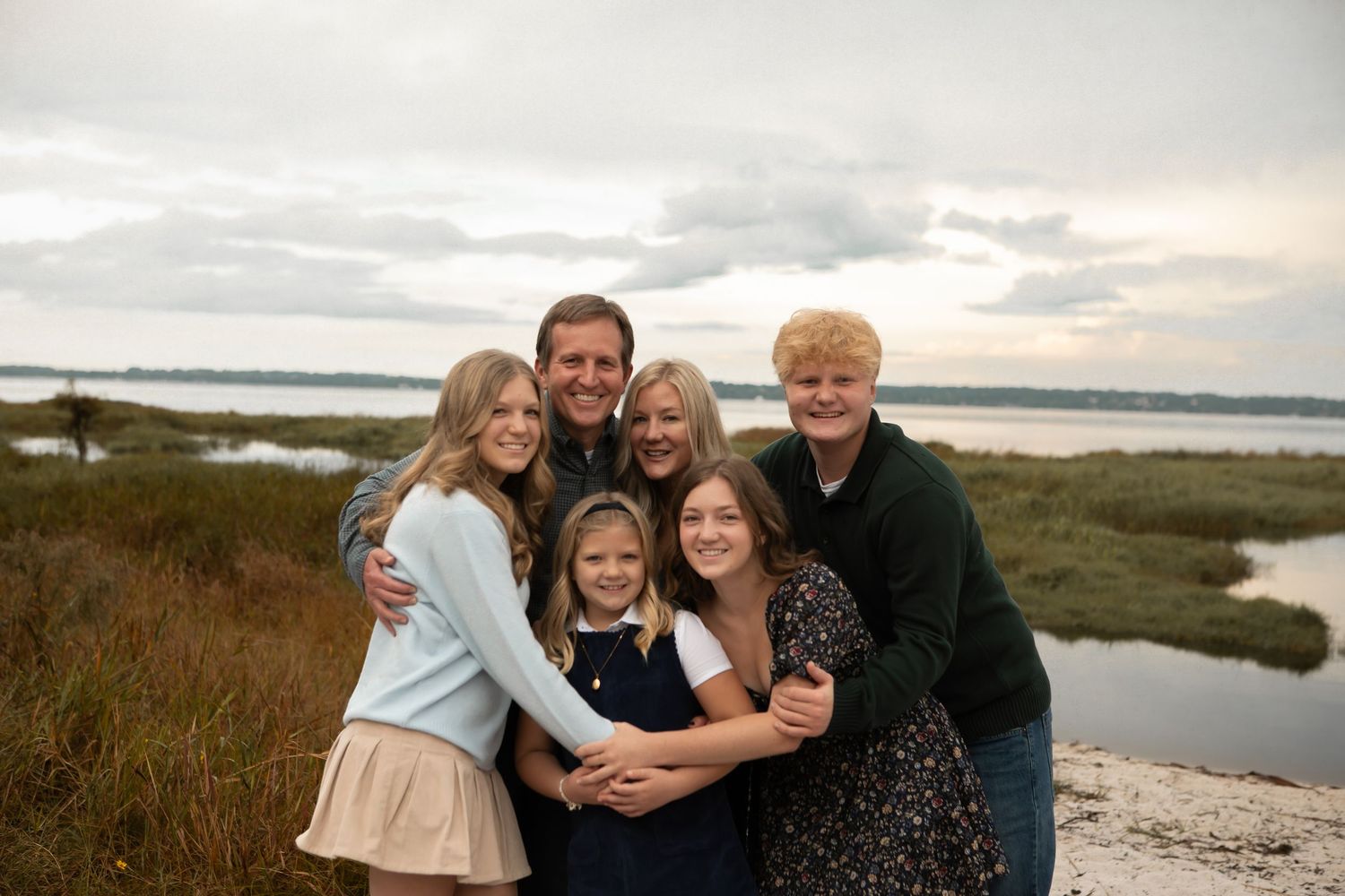 A smiling family of six embraces closely outdoors near a marsh under a cloudy sky.