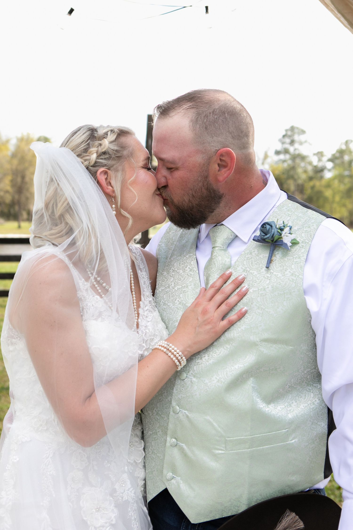 Bride and groom share their first kiss, with bride in lace gown and groom in mint vest at wedding.