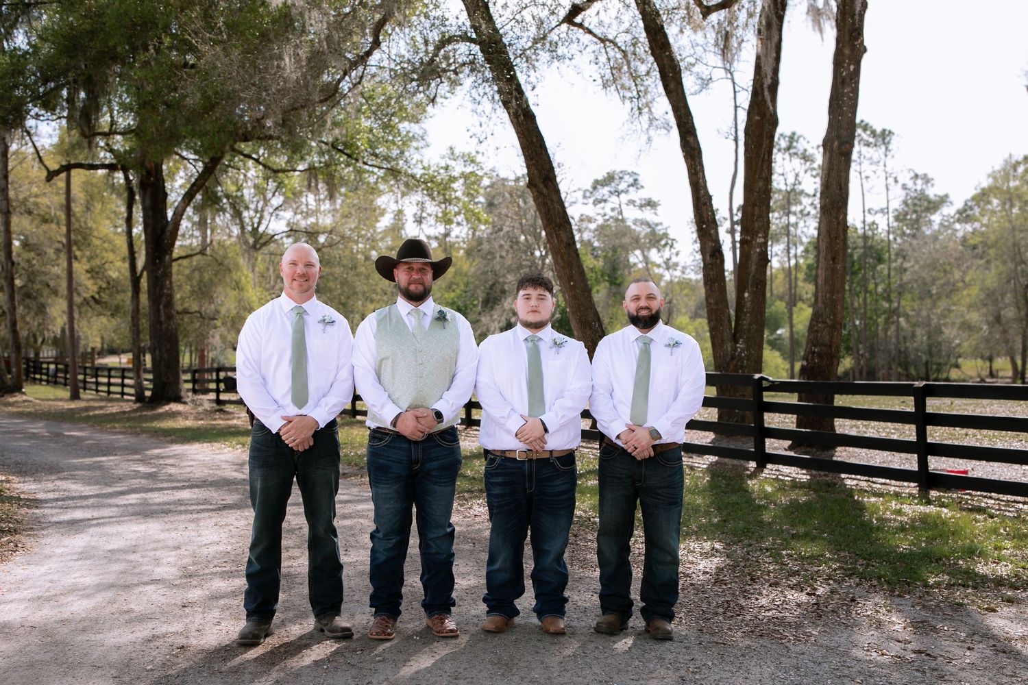 Groom and three groomsmen in white shirts, sage ties, and jeans posing at rustic ranch venue.