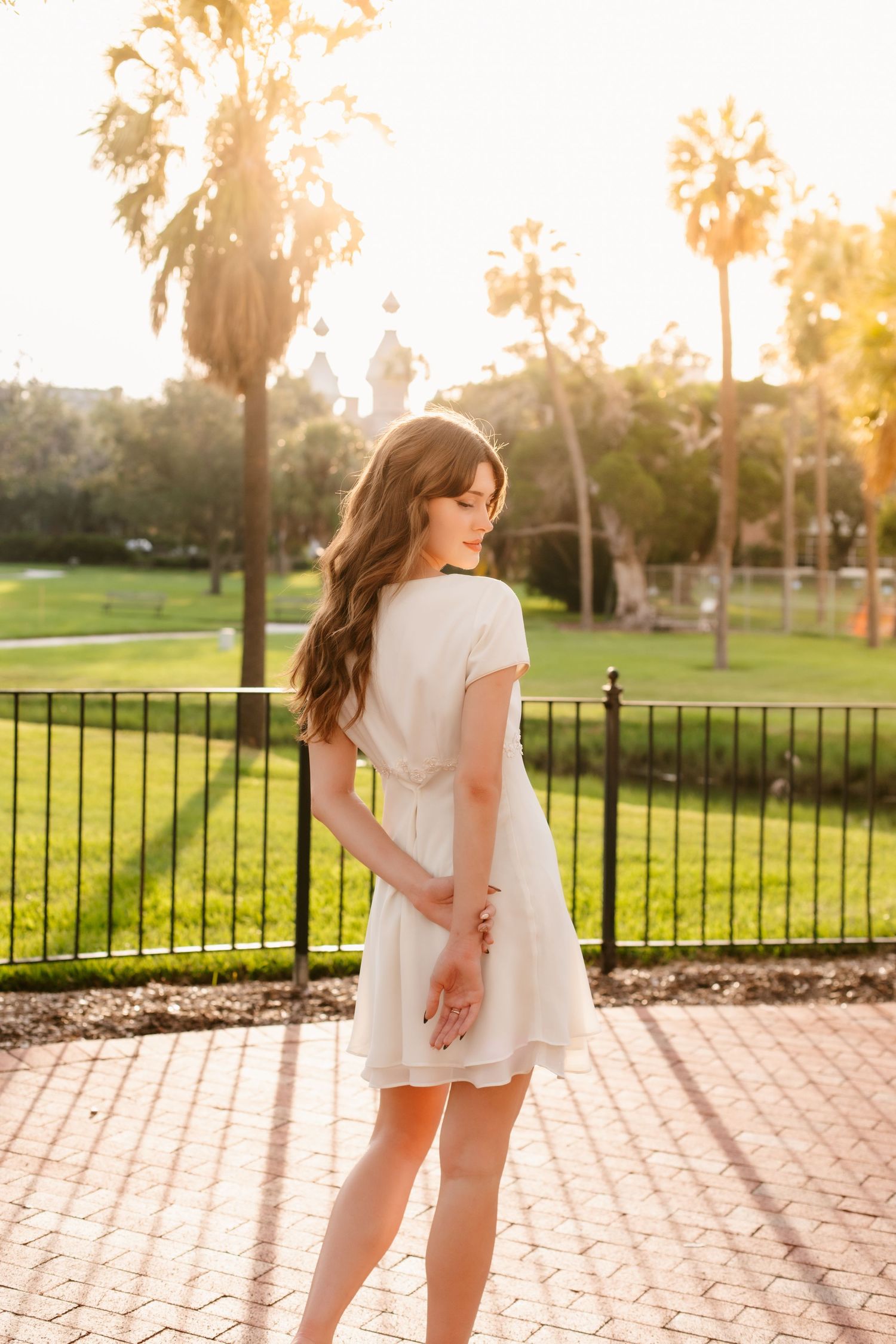 Young woman in white dress standing in sunny park with palm trees, golden hour light glowing behind her.