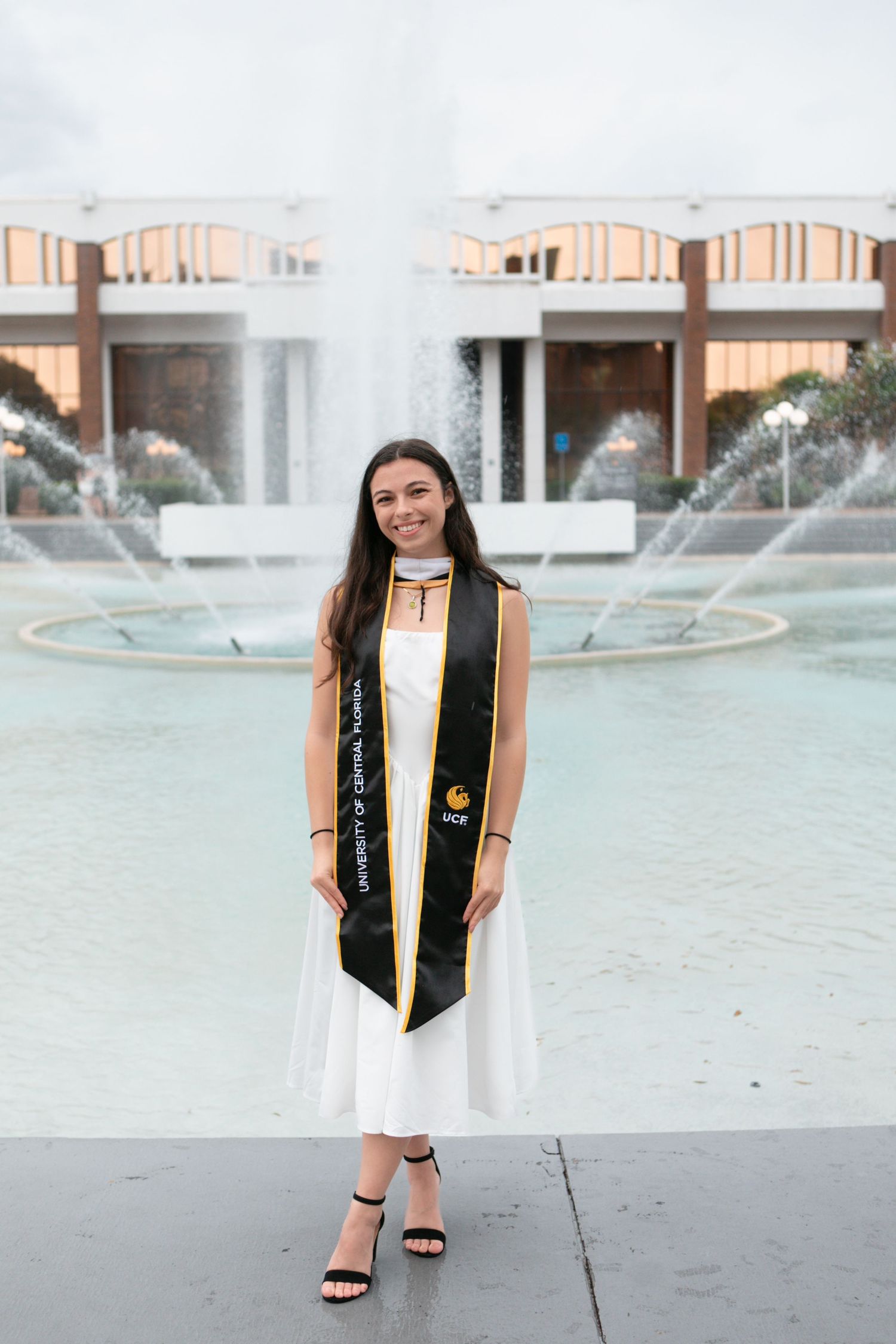 Graduate proudly wearing University of Central Florida sash stands before campus fountain.