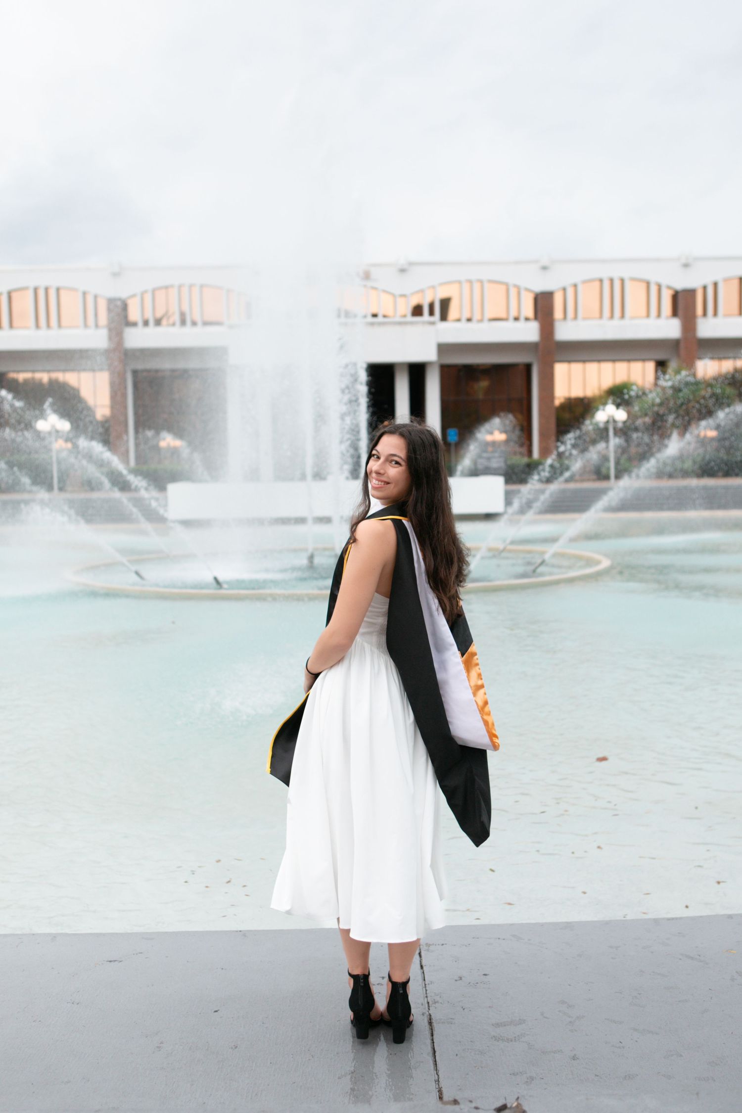 UCF graduate in white dress and black sash poses by campus fountain.