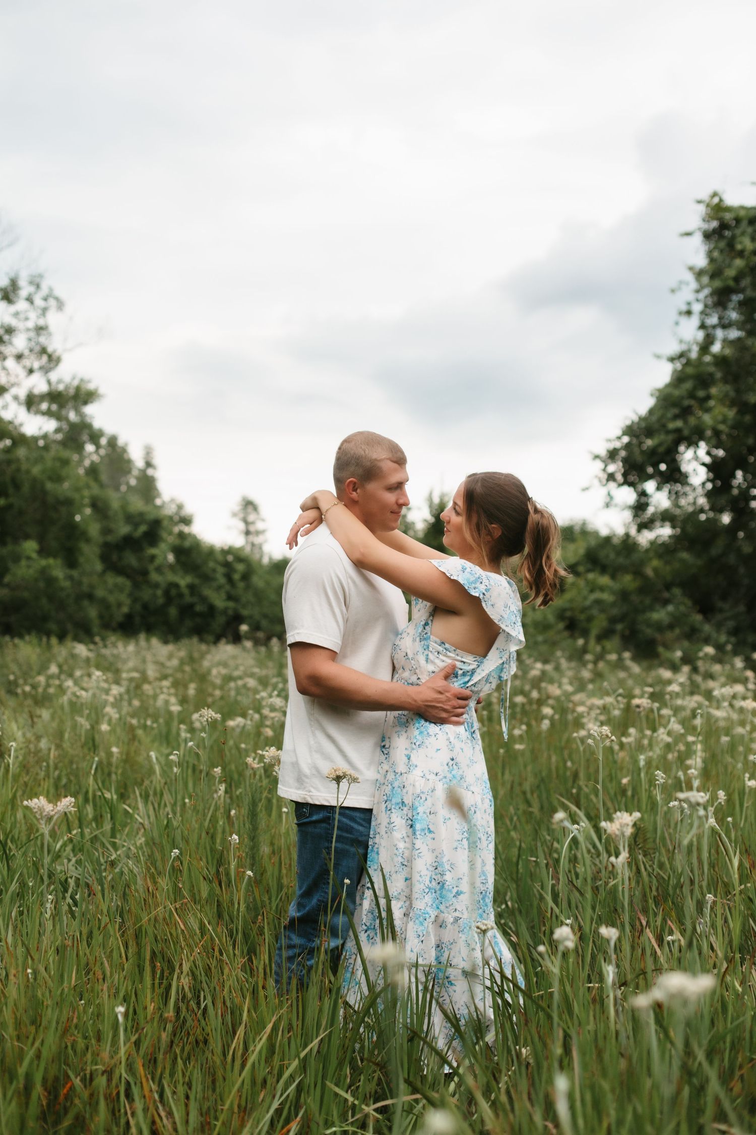 Couple embracing in a wildflower field, woman in floral dress and man in white shirt, surrounded by green trees.