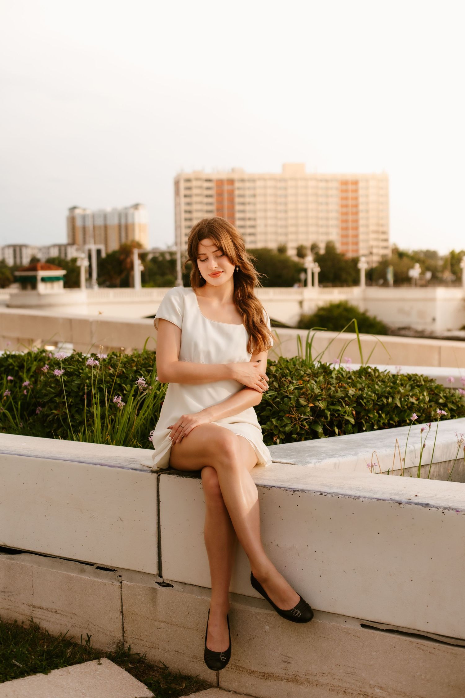 Young woman in white dress sitting on a concrete ledge with city buildings and greenery in the background.
