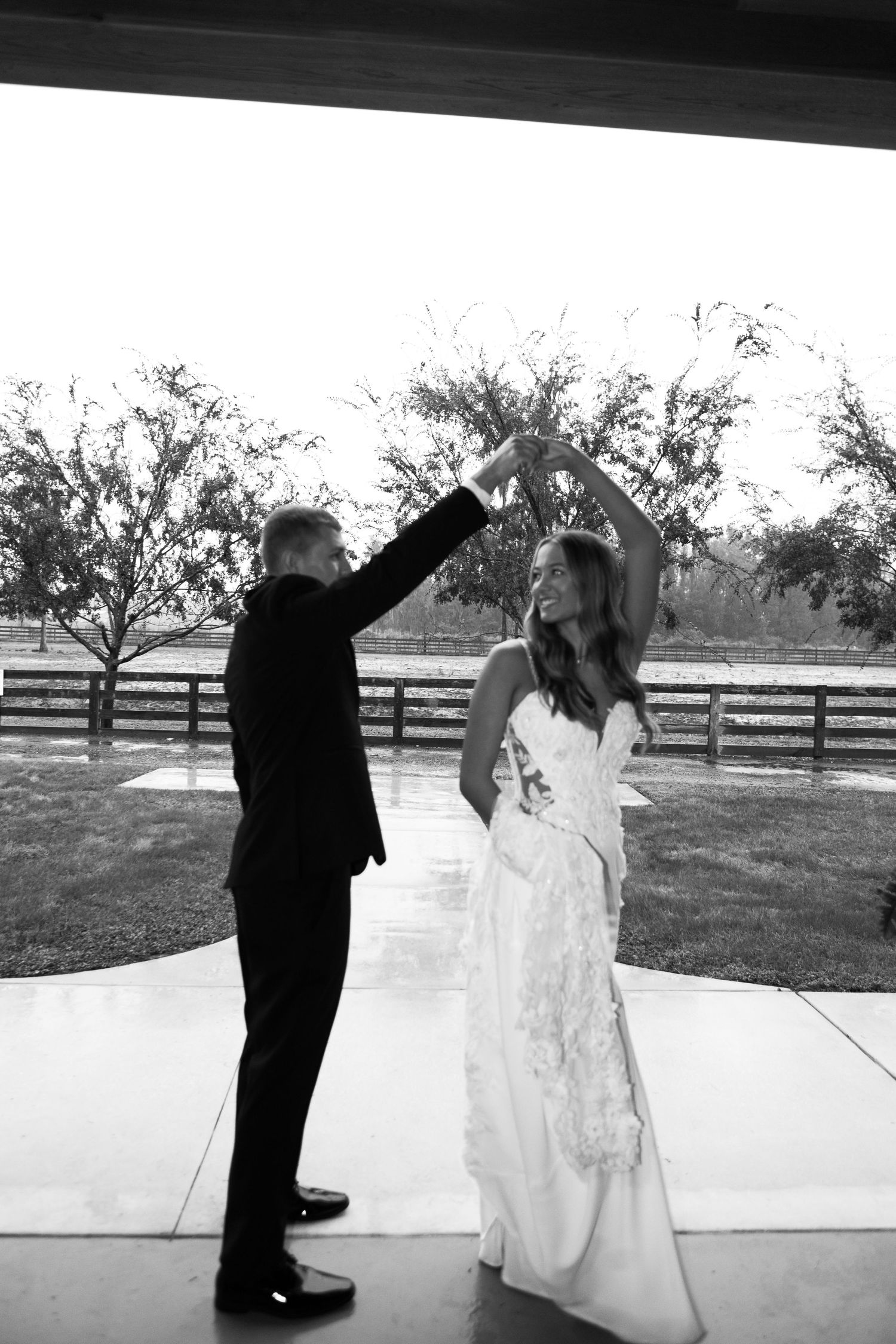 Black and white photo of a groom spinning his bride in a wedding dance outdoors near a rustic fence.