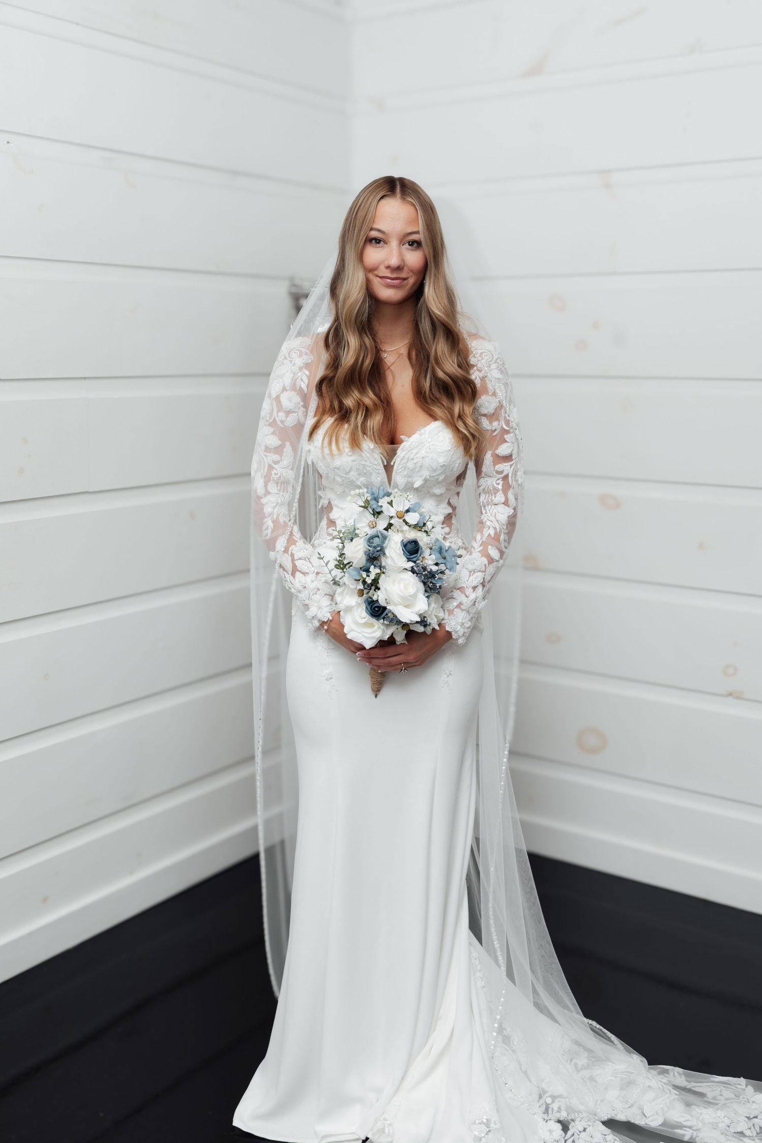Bride in lace long-sleeve wedding gown with cathedral veil holds white and blue bouquet indoors.
