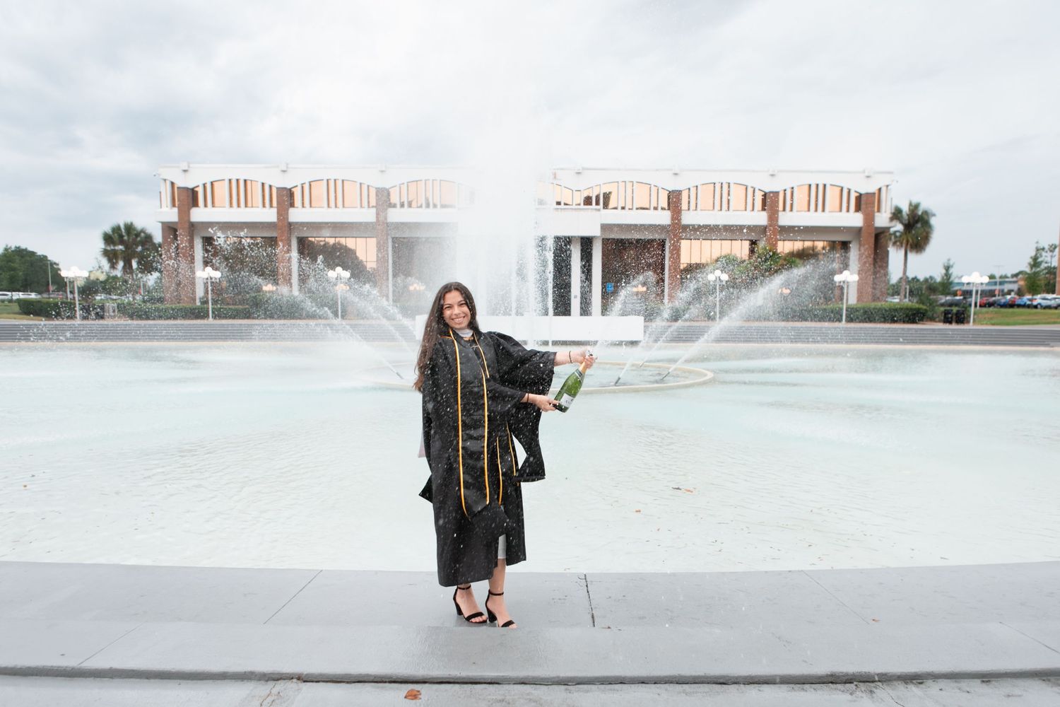 Graduate in black cap and gown poses proudly in front of a fountain at UCF campus.