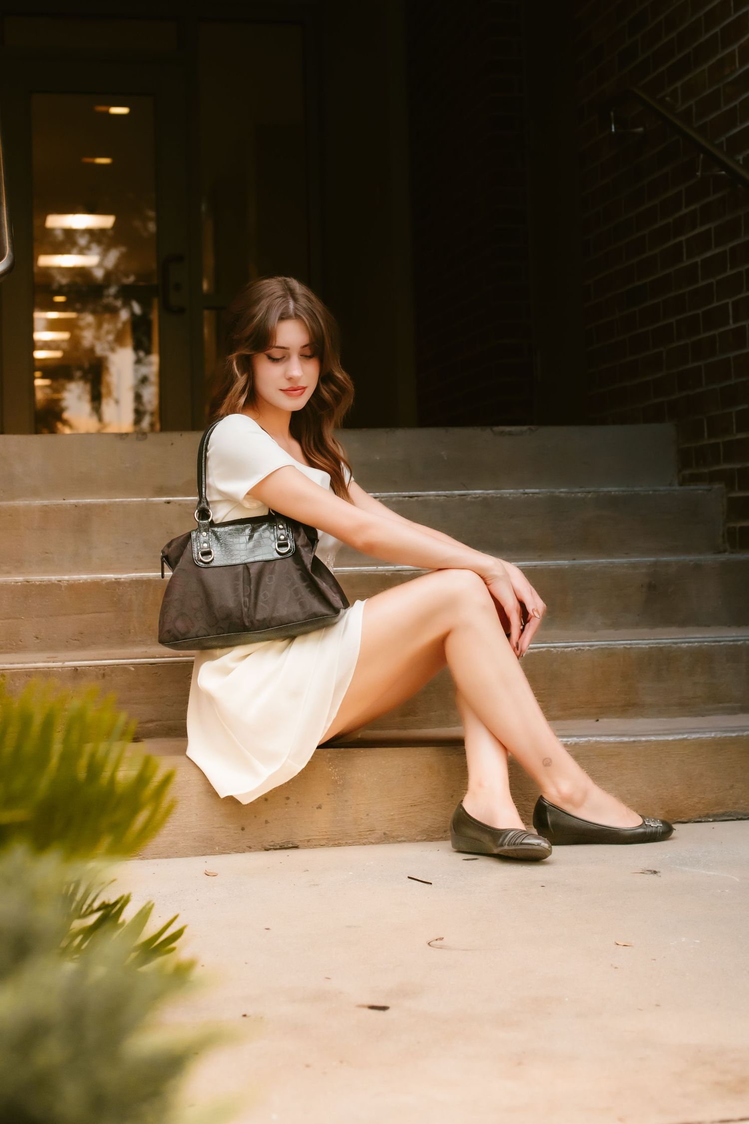 Young woman in white dress and black flats sitting on stone steps with leather handbag.