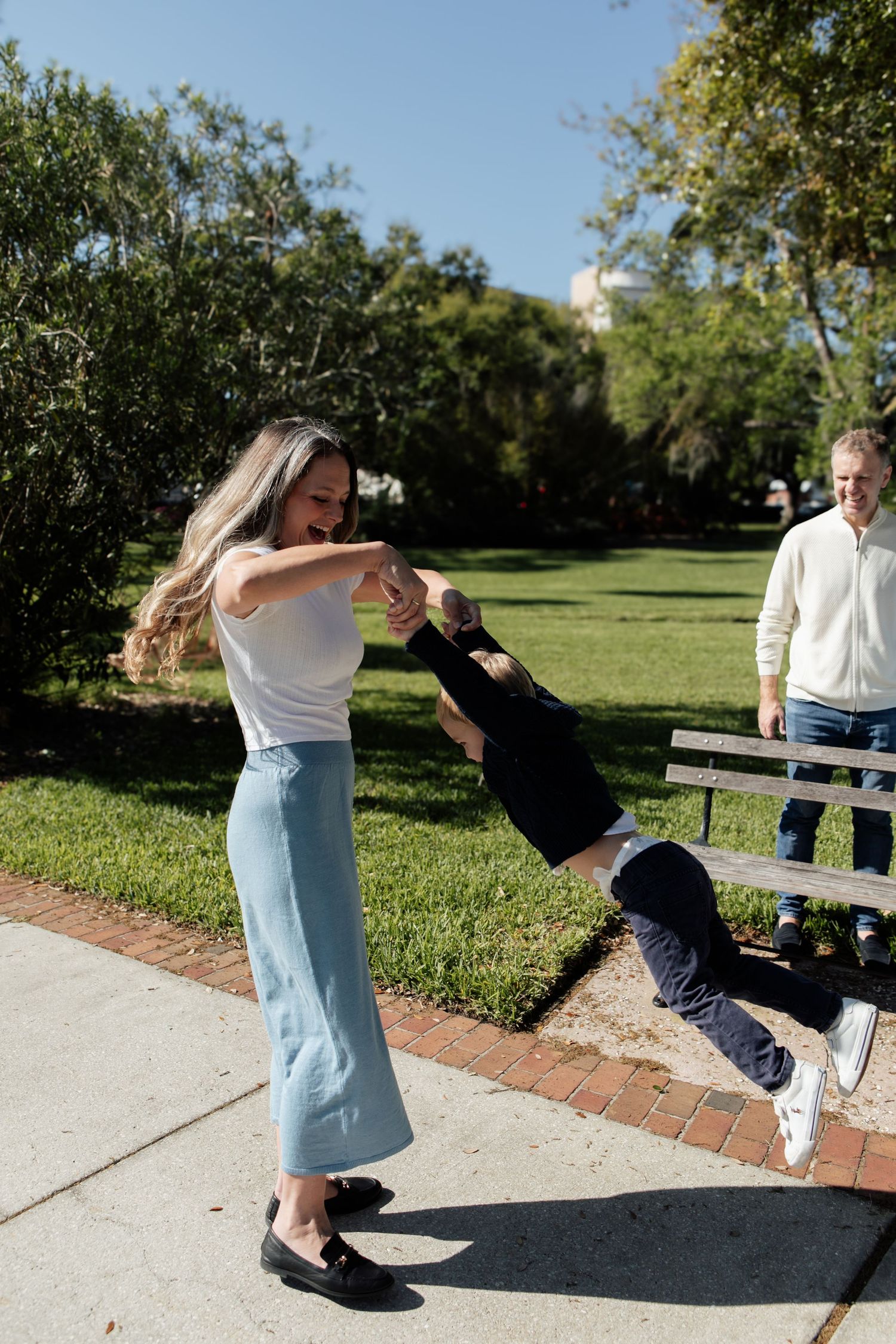 A mother joyfully spins her child in a sunny park while a man watches from a bench.