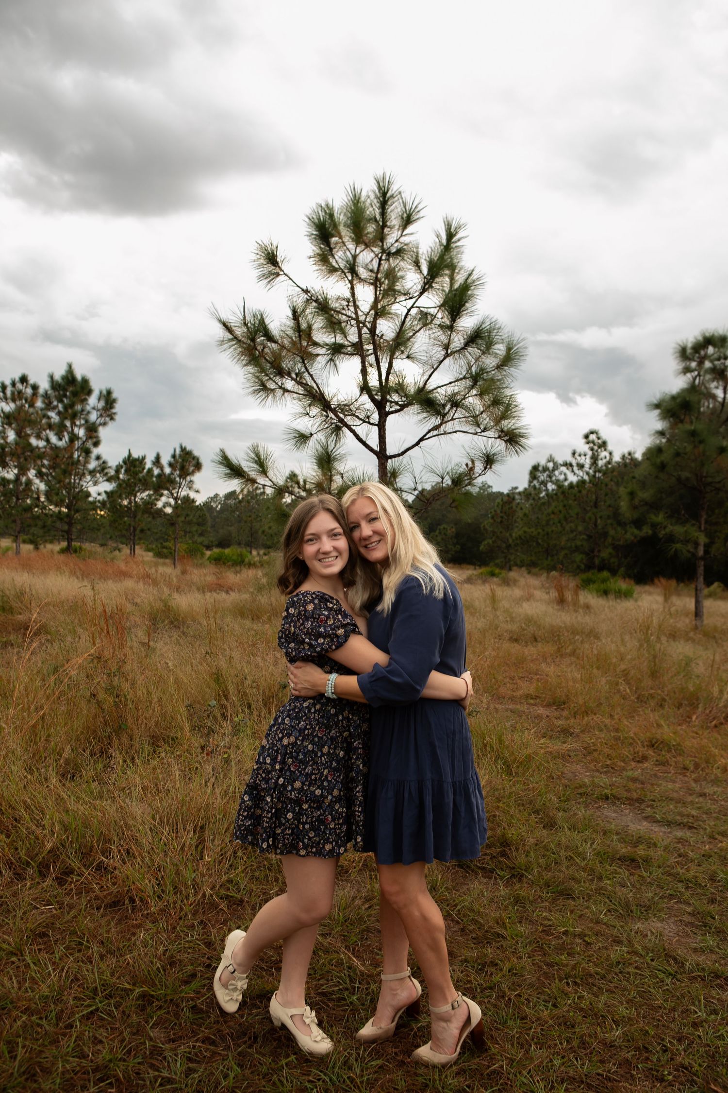 Two young women in dresses hugging and smiling outdoors among pine trees on a cloudy day.