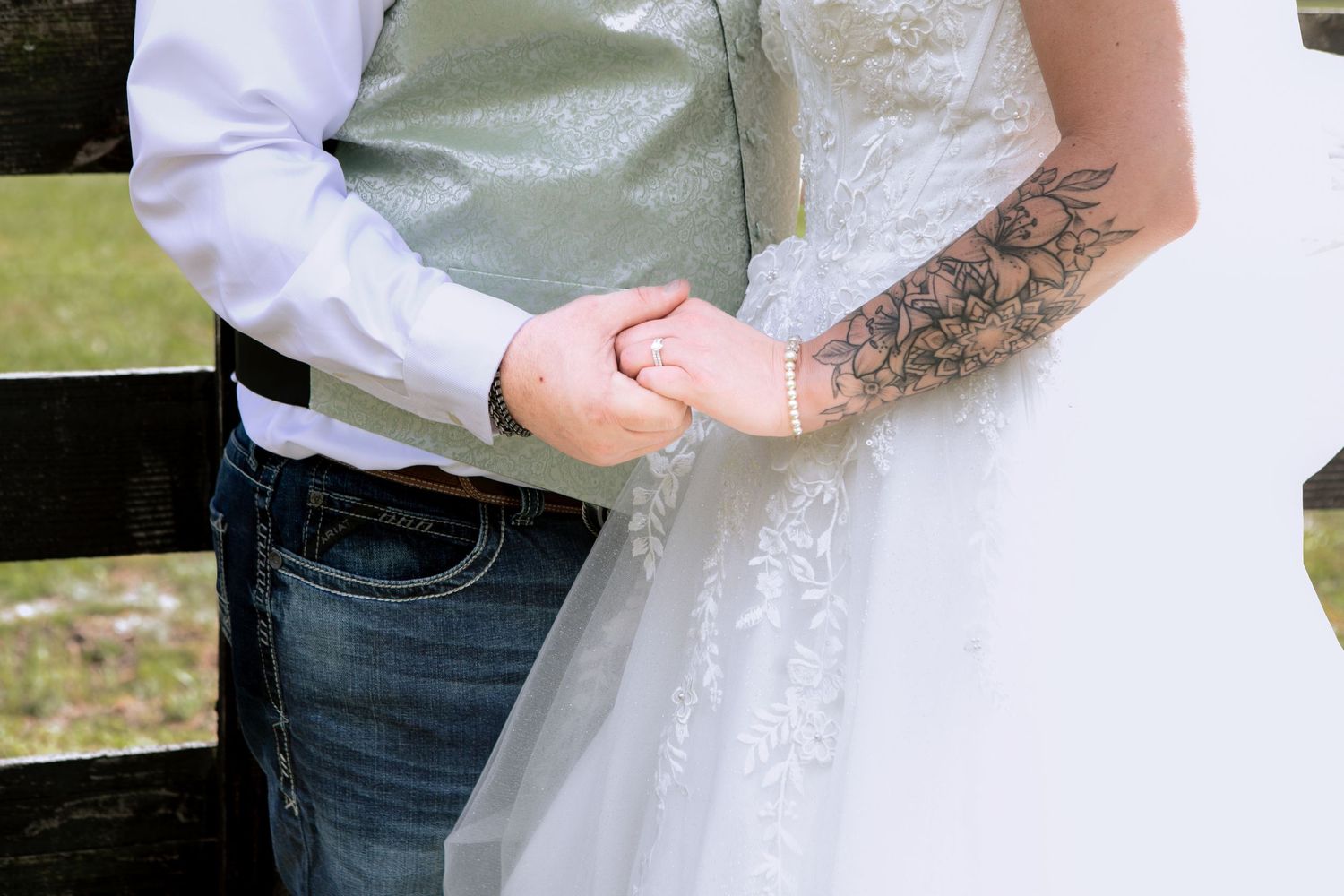 Bride in lace wedding dress with floral tattoo holding hands with groom in jeans near wooden fence.