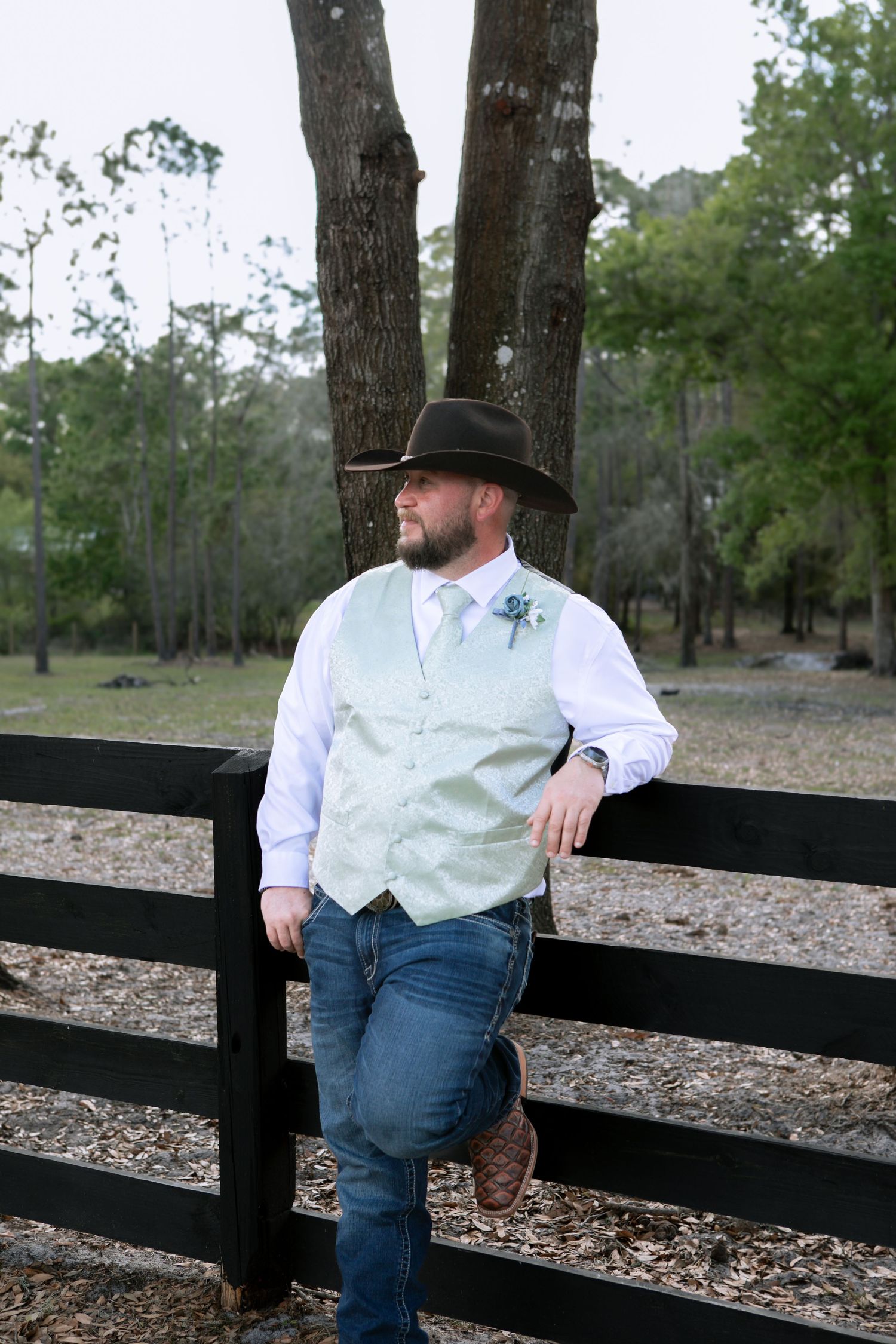 Bearded groom wearing cowboy hat and mint vest with boutonniere poses by black fence at rustic farm wedding.