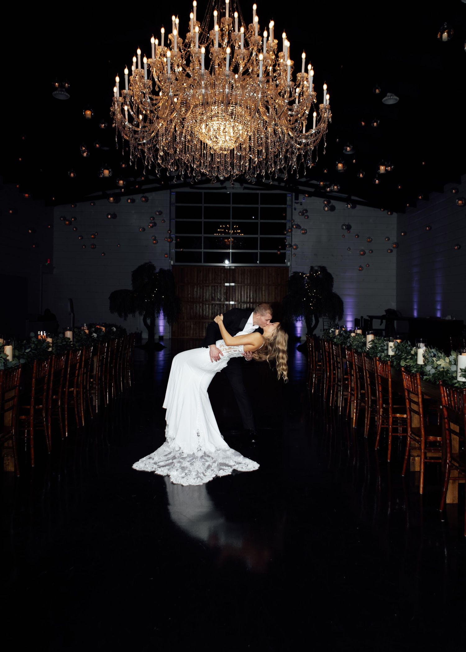 Bride and groom share a dramatic dip kiss under a grand chandelier in a dark wedding venue.