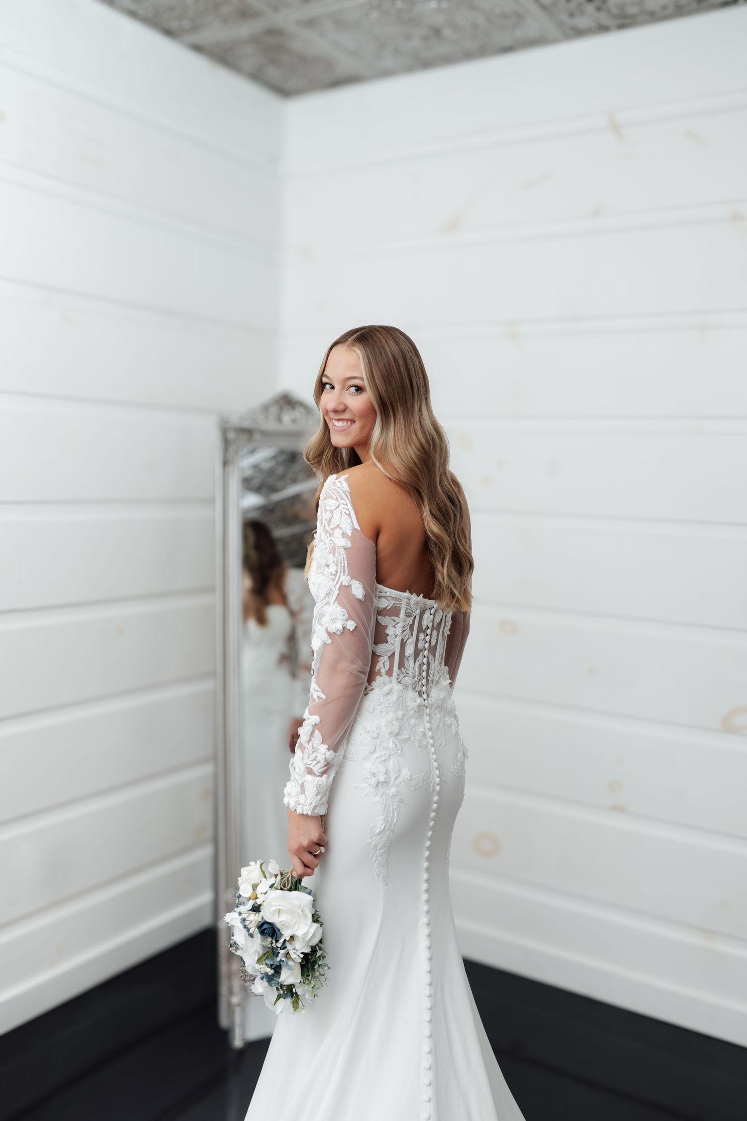 Smiling bride in lace long-sleeve backless wedding gown holds bouquet while posing near ornate mirror.
