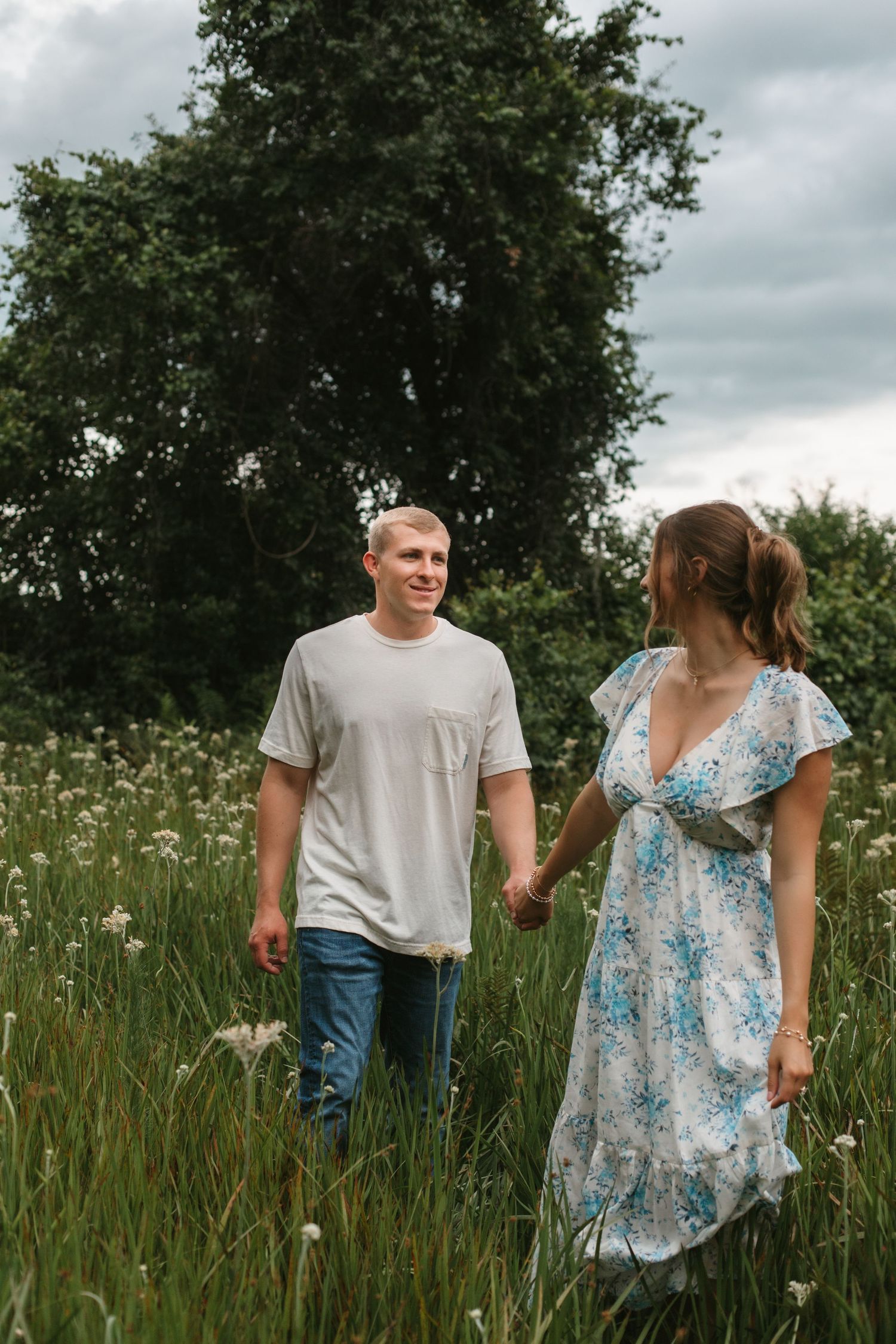 A couple holds hands walking through a lush green meadow with wildflowers, smiling at each other on a cloudy day.