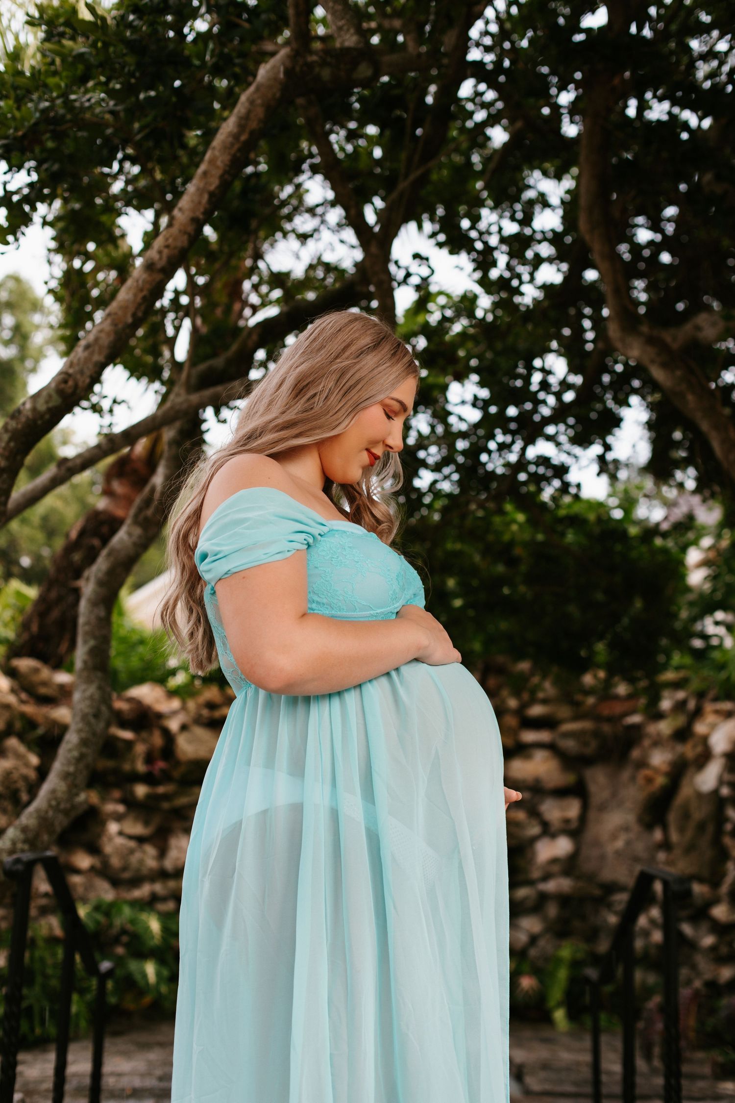 Pregnant woman in a light blue off-shoulder gown cradling her baby bump outdoors among lush green trees.
