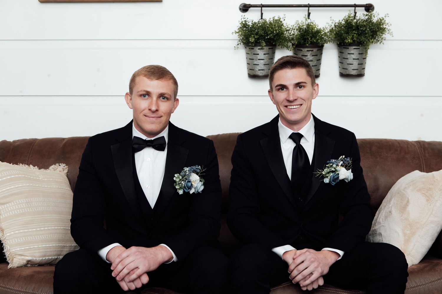 Two grooms in black tuxedos with boutonnieres sit together on a brown leather couch at their wedding.