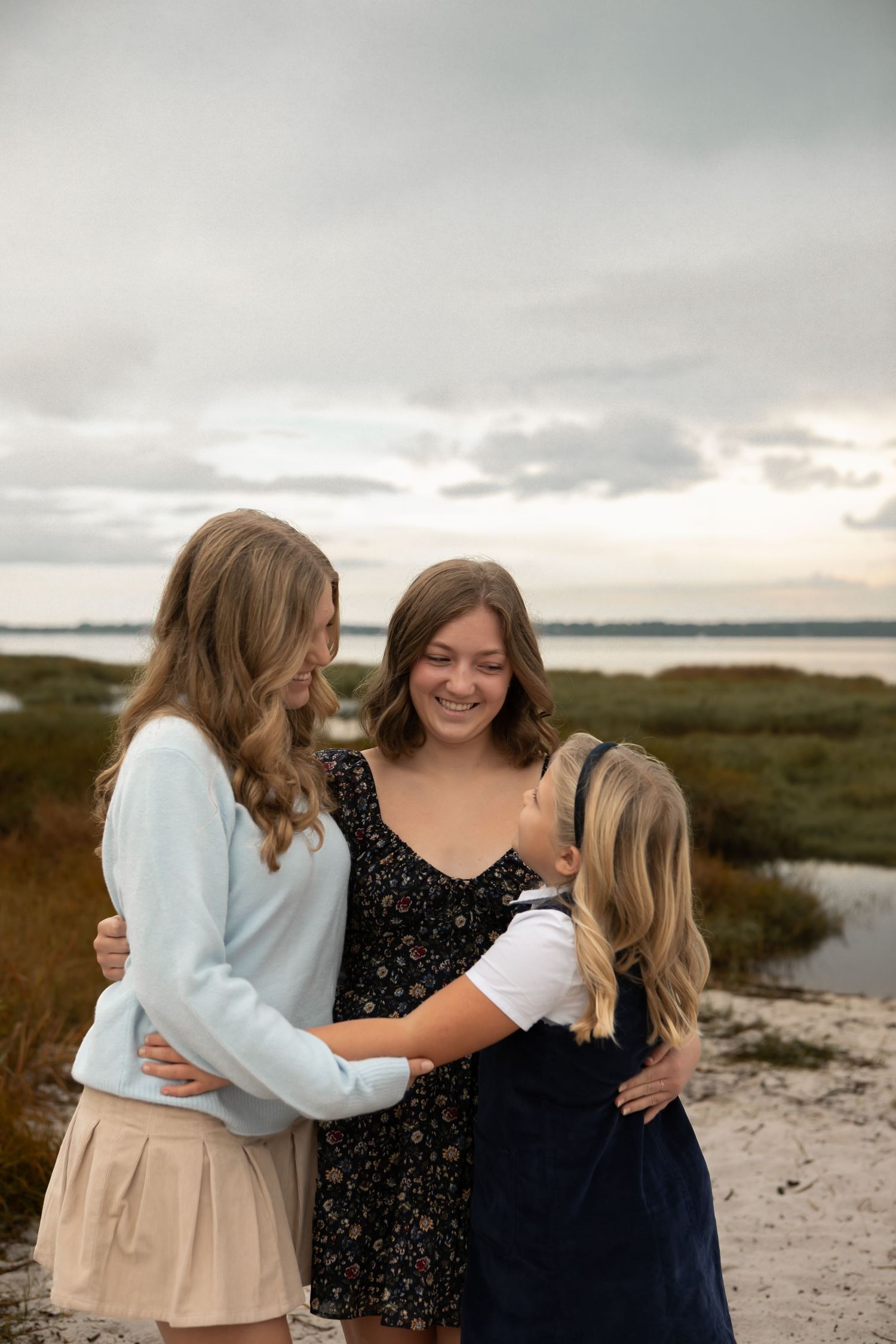 Three teenage girls hugging and smiling together on a sandy beach with marshland and cloudy sky behind them.