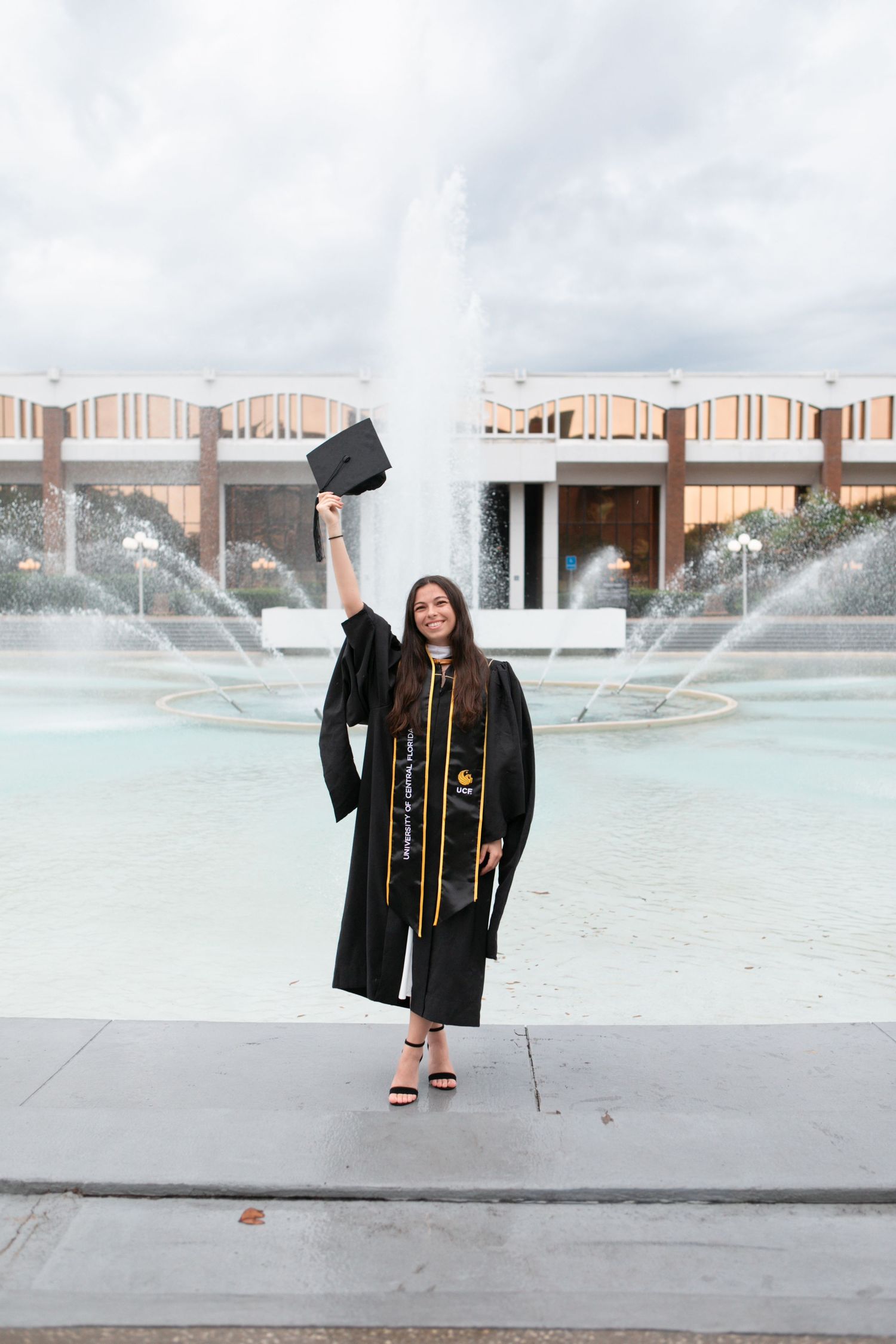 Joyful female graduate raising cap in the air while standing in front of UCF fountain.