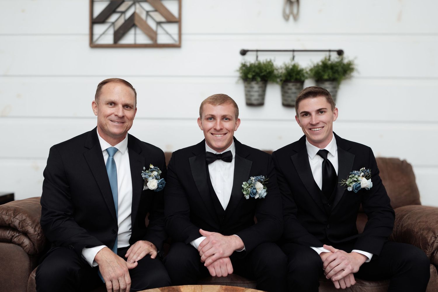 Three men in black suits with boutonnieres smile while seated on a leather couch at a wedding venue.