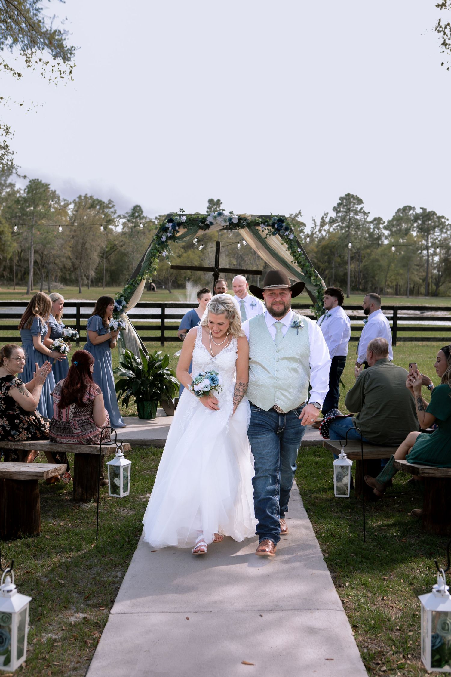 Bride and groom walk down outdoor wedding aisle, surrounded by guests, with a floral arch and green pasture backdrop.