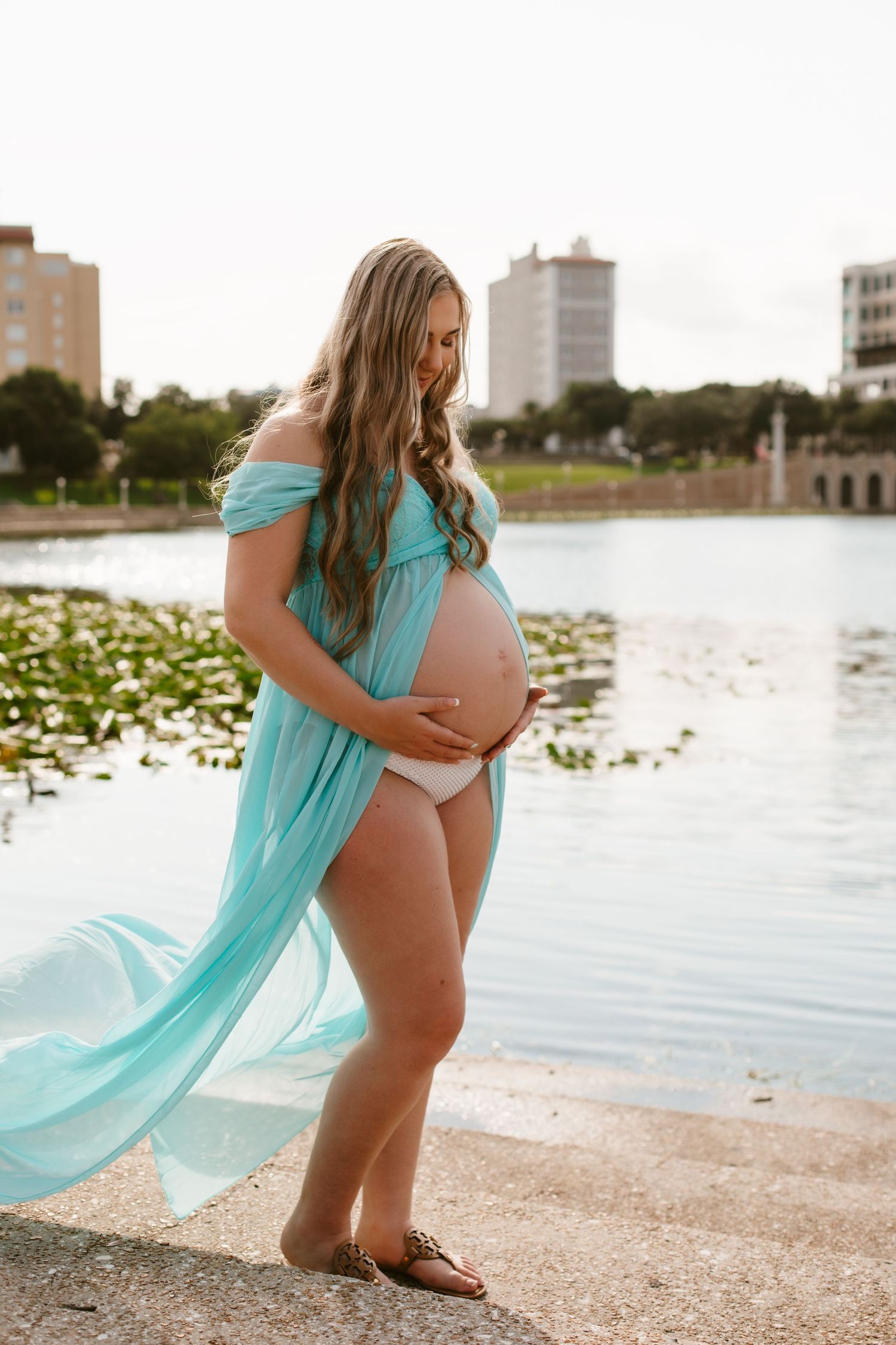 Pregnant woman in flowing teal off-shoulder gown cradling her baby bump by a serene lakeside.
