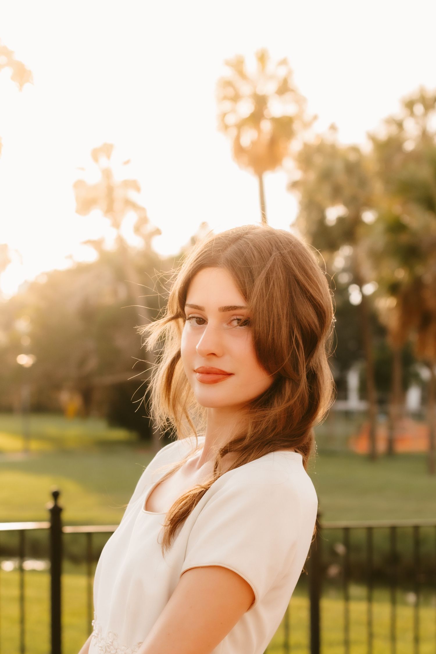 Young woman with brown hair in white dress poses outdoors at golden hour with palm trees in background.