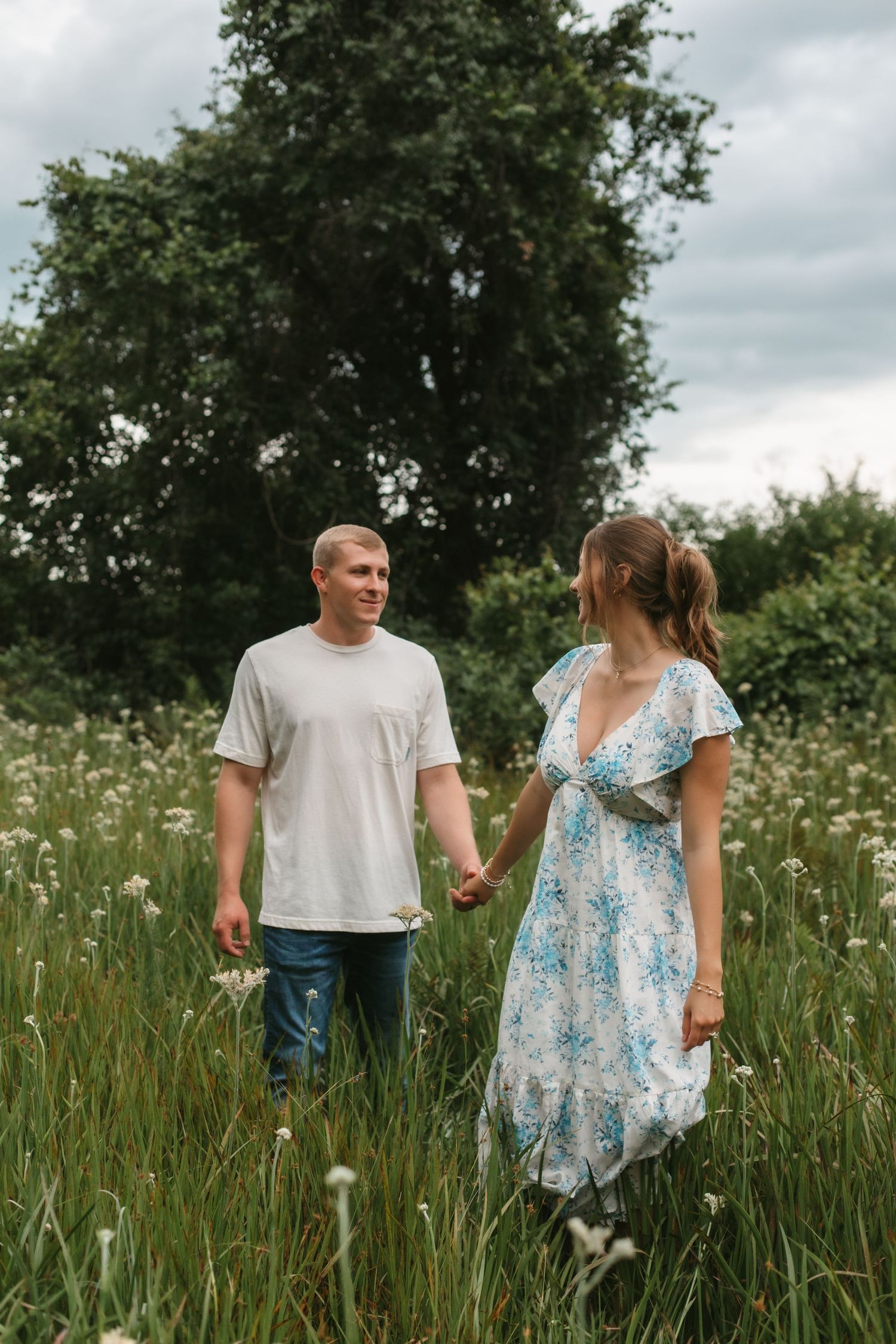 Couple holding hands in a wildflower meadow, man in white tee and jeans, woman in floral dress, green trees behind.
