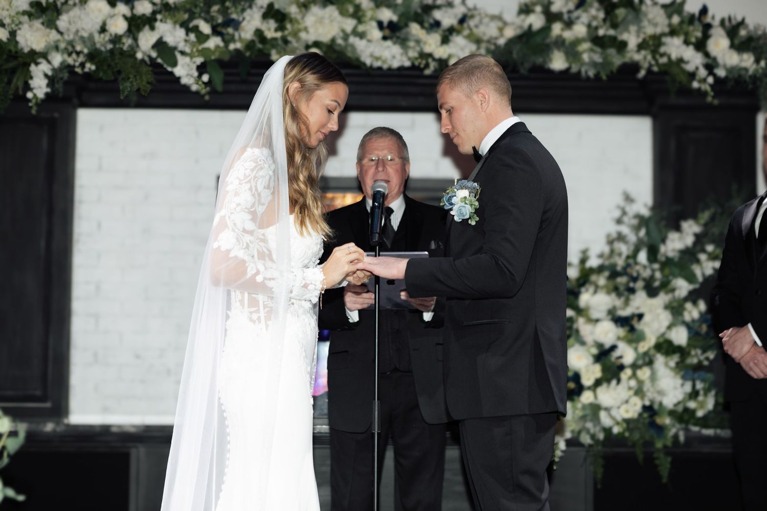 Bride and groom exchanging rings at elegant wedding ceremony with white floral arrangements and officiant in background.