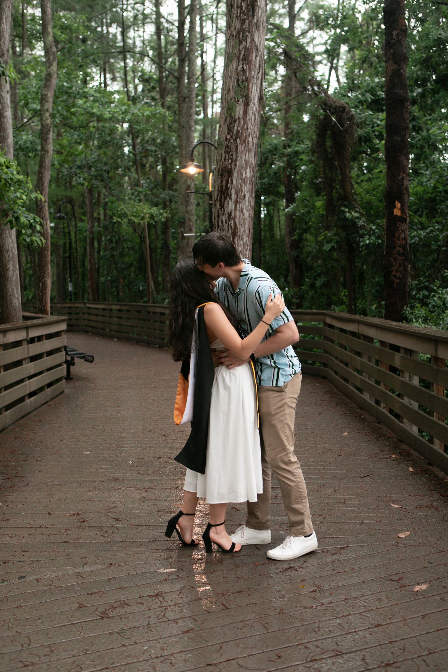Couple embraces and kisses on wooden bridge surrounded by lush green forest after graduation.