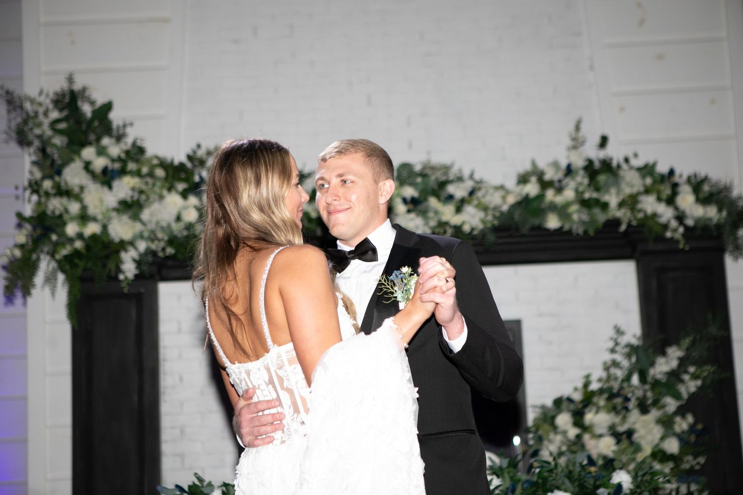 Newlywed couple sharing first dance at elegant wedding reception with white floral backdrop.