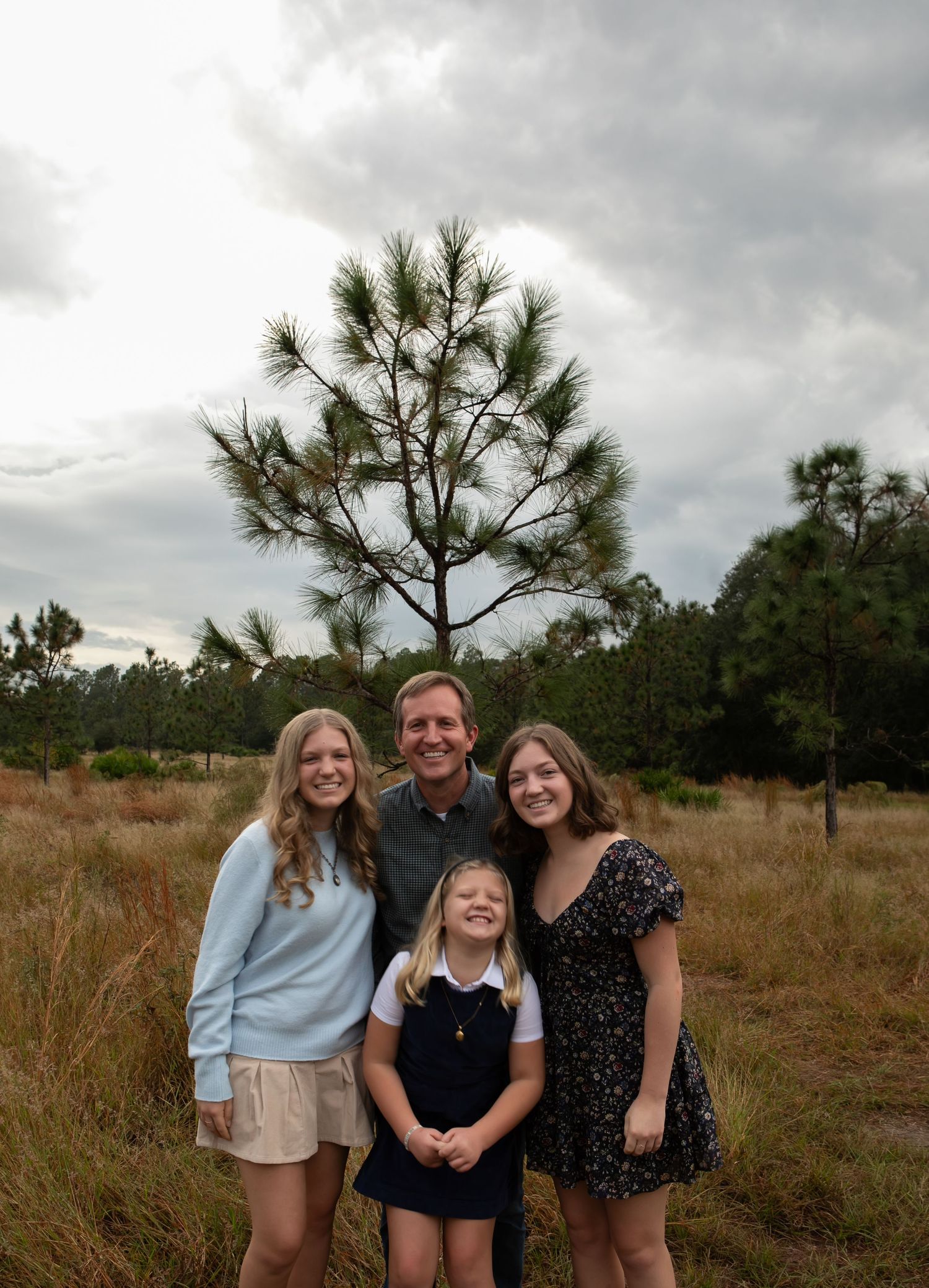 A family of four poses outdoors in a grassy field with pine trees and cloudy sky backdrop.