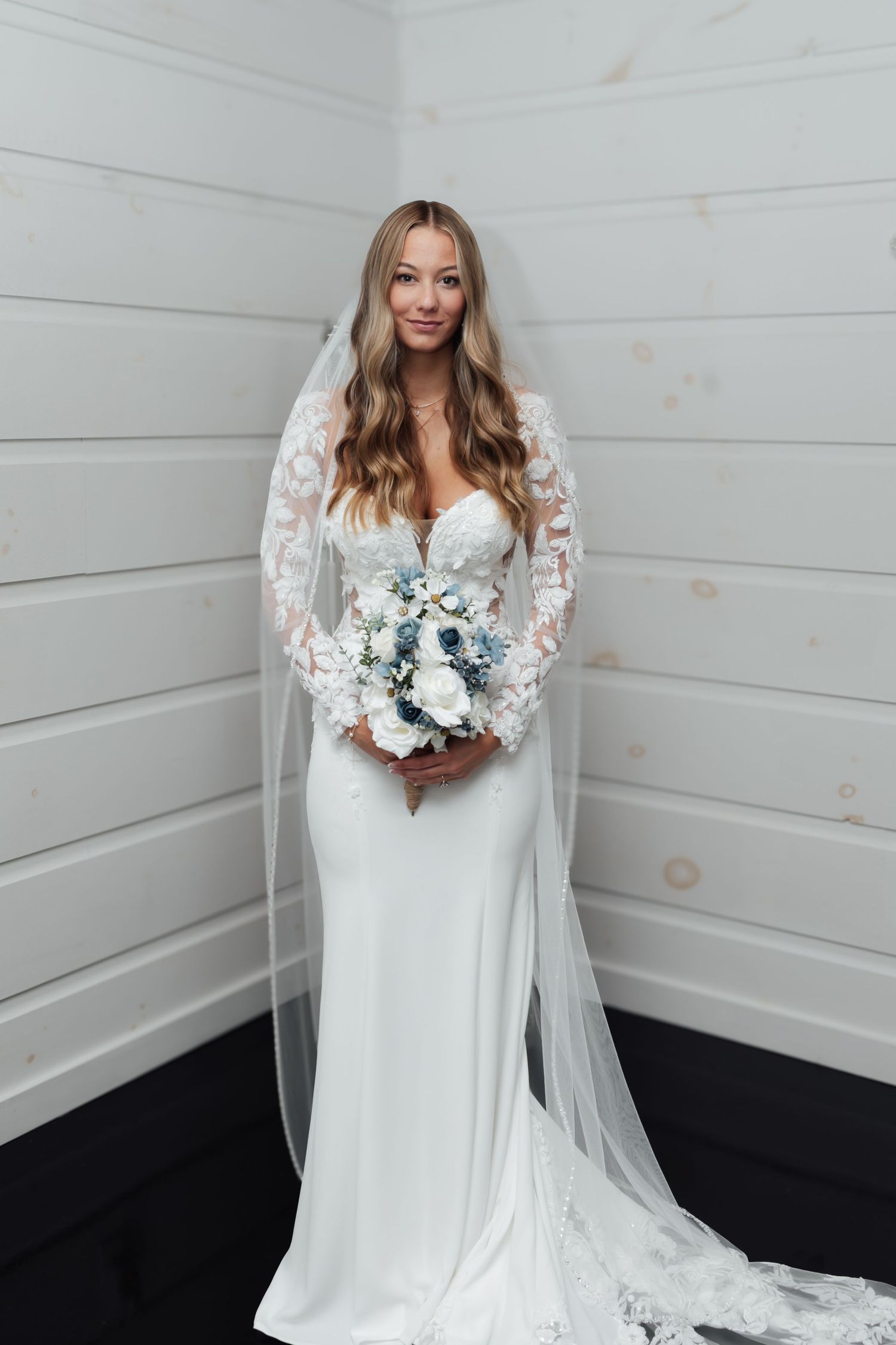 Bride in a white lace-sleeve gown holding a blue and white bouquet with a cathedral veil.