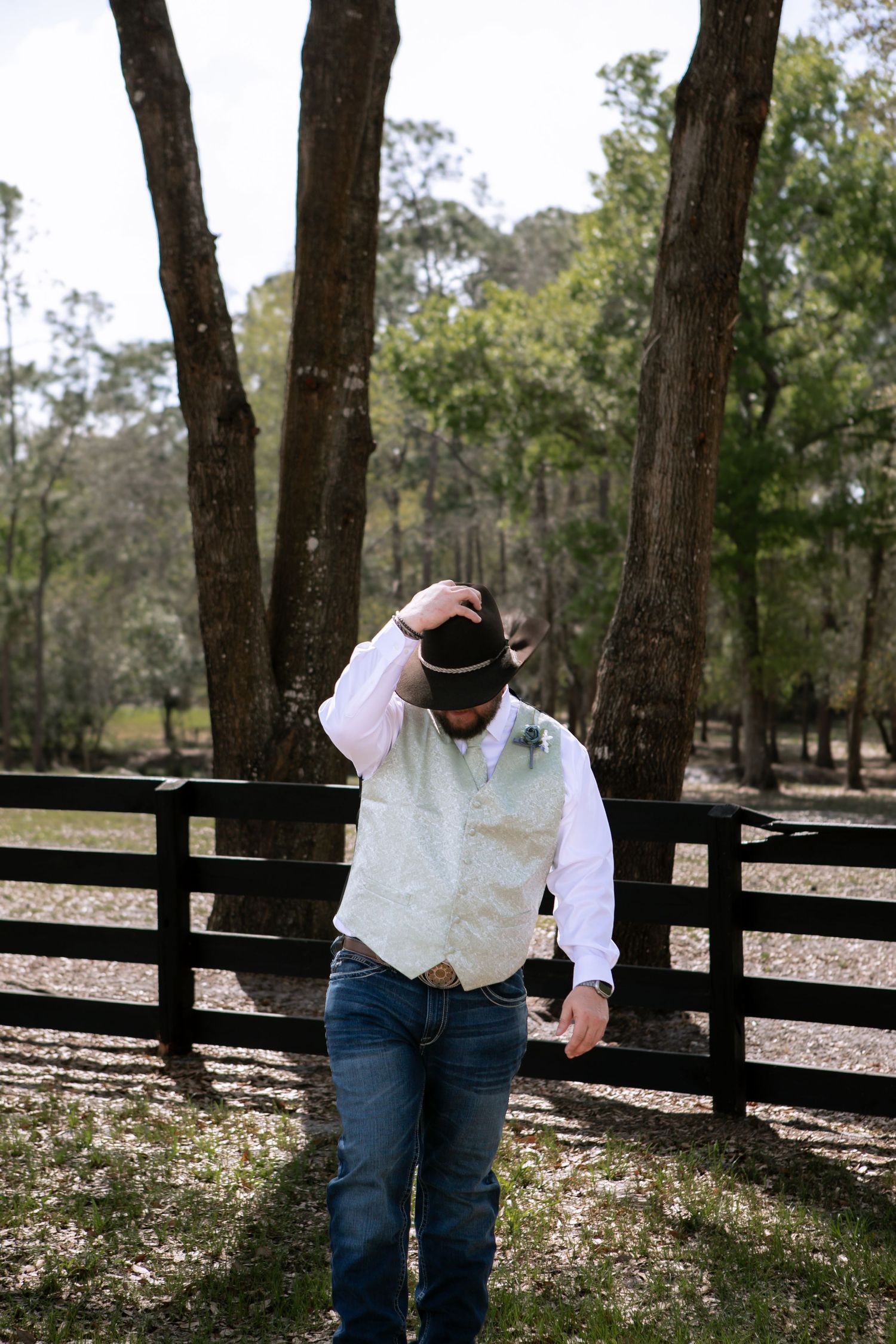 Groom in white vest, jeans, and cowboy hat adjusting his hat outdoors near a wooden fence at a rustic wedding.