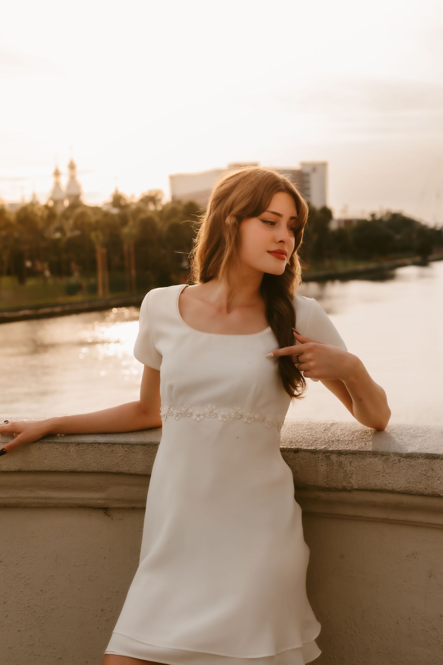 Young woman in white mini dress poses by stone railing at golden hour with river and city skyline behind her.