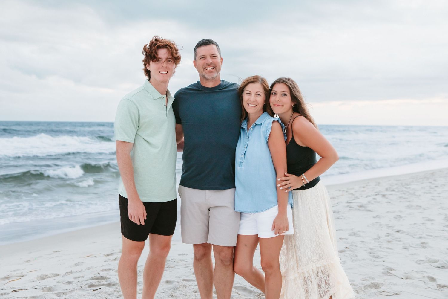 Family of four smiling together on a sandy beach with ocean waves in the background.
