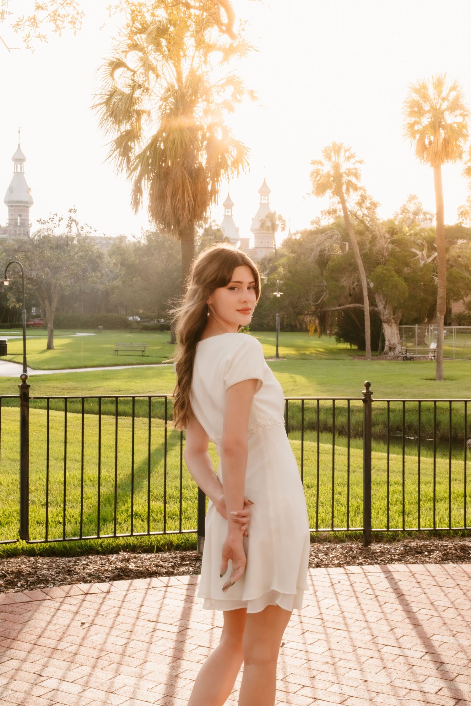 Young woman in white dress poses by iron fence at golden hour with palm trees and historic building backdrop.