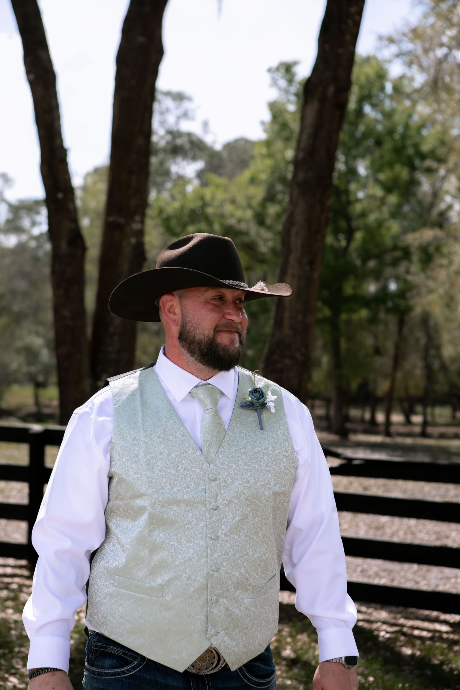 Handsome groom in cowboy hat, sage vest, and boutonniere posing at rustic outdoor ranch wedding.